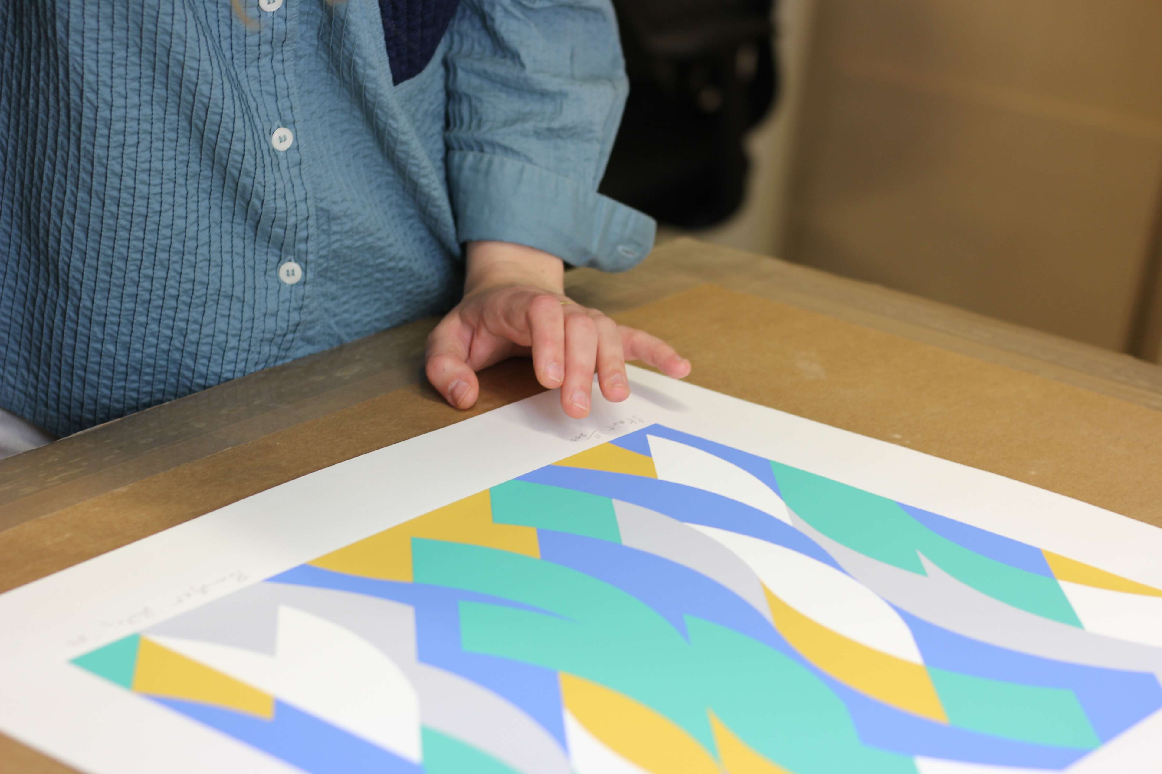 A person pointing to an artwork on a table containing a pattern of unusual shapes in green, blue, yellow, grey, and white.