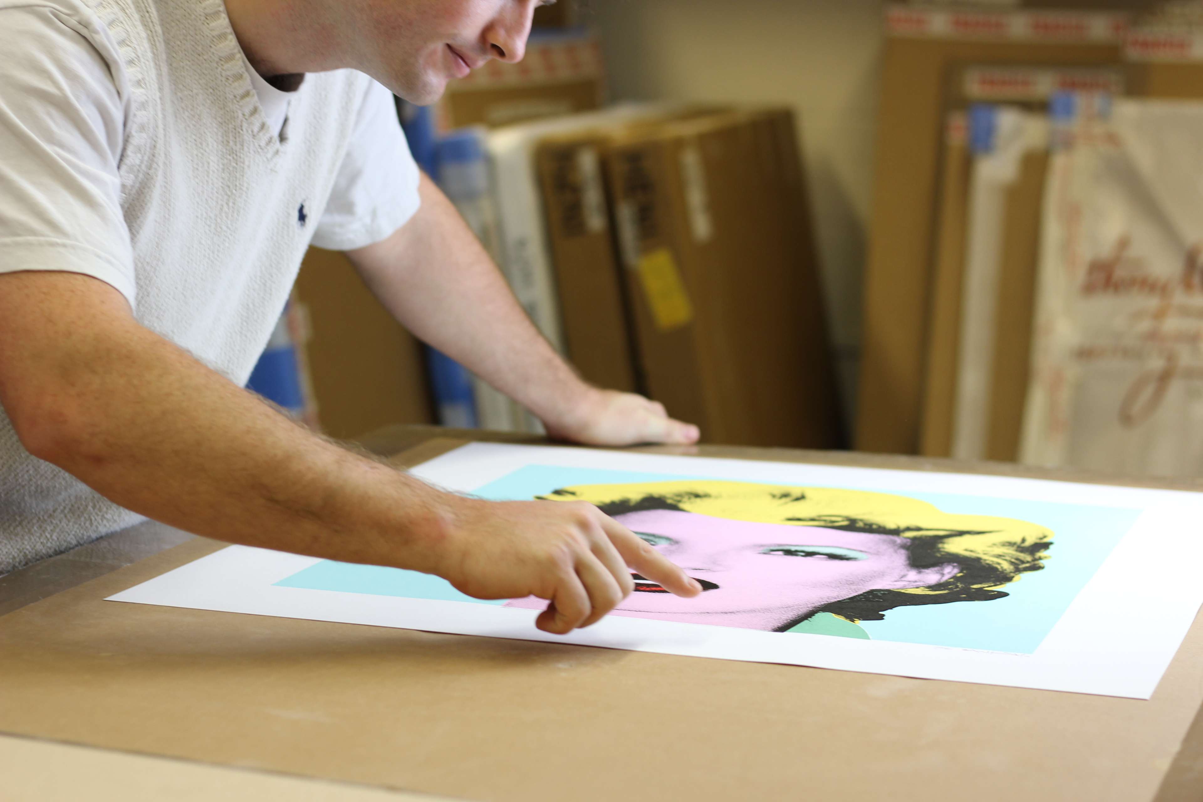 A man leaning over an artwork on a table and pointing at the print, which depicts Kate Moss in the likeness of Kate Moss with bright yellow hair against a mint green background.