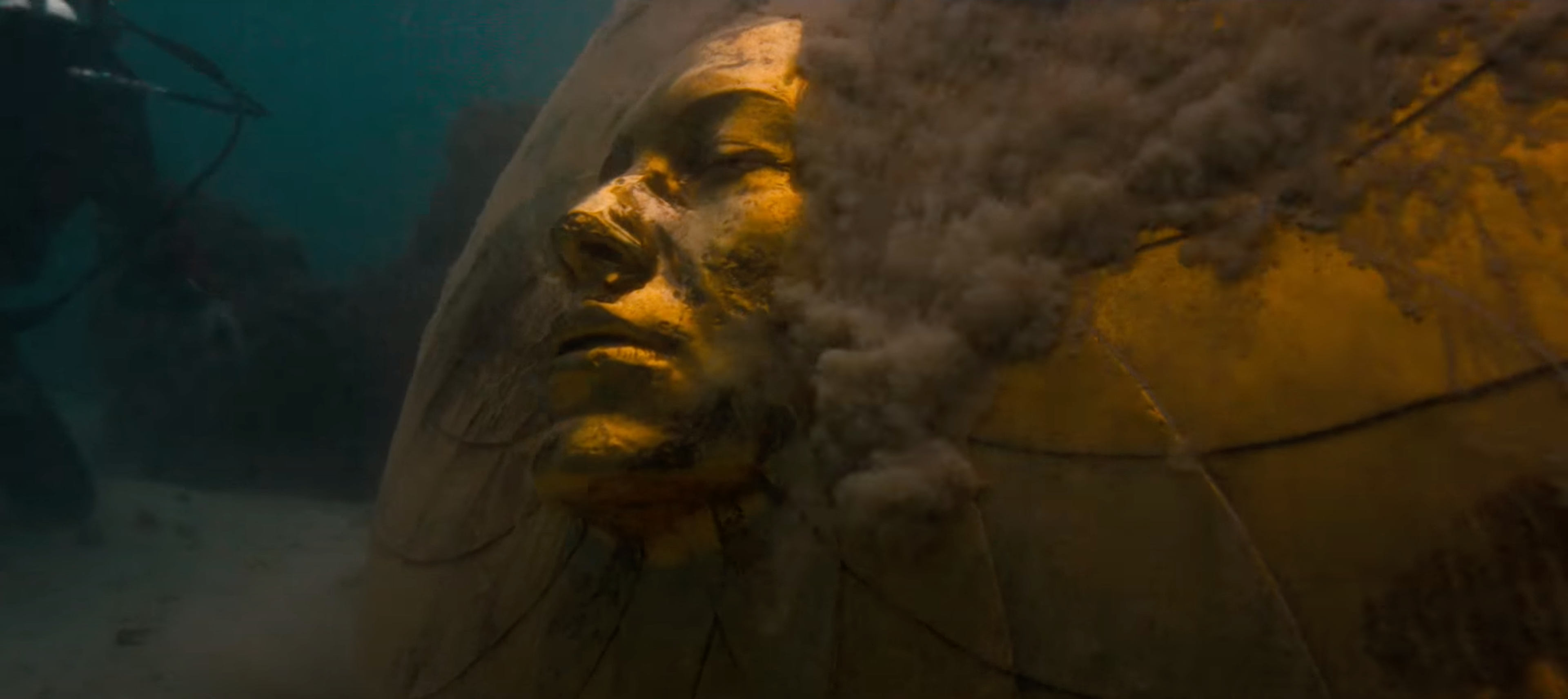 A large-scale golden sculpture depicting a face is seen underwater, surrounded by sand.