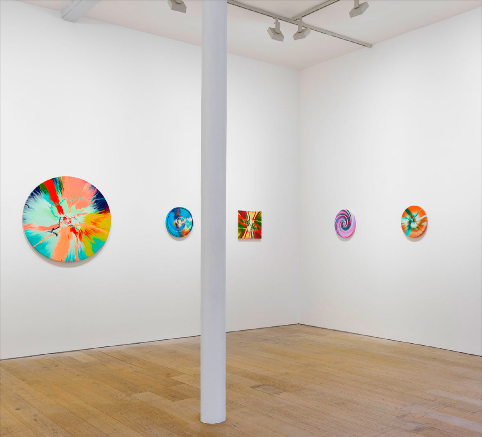 Installation view of Damien Hirst's The Beautiful Paintings in a gallery room. The white gallery wall has a simple hang of the works, featuring four circular compositions and one square work, in varying colour palettes and sizes.