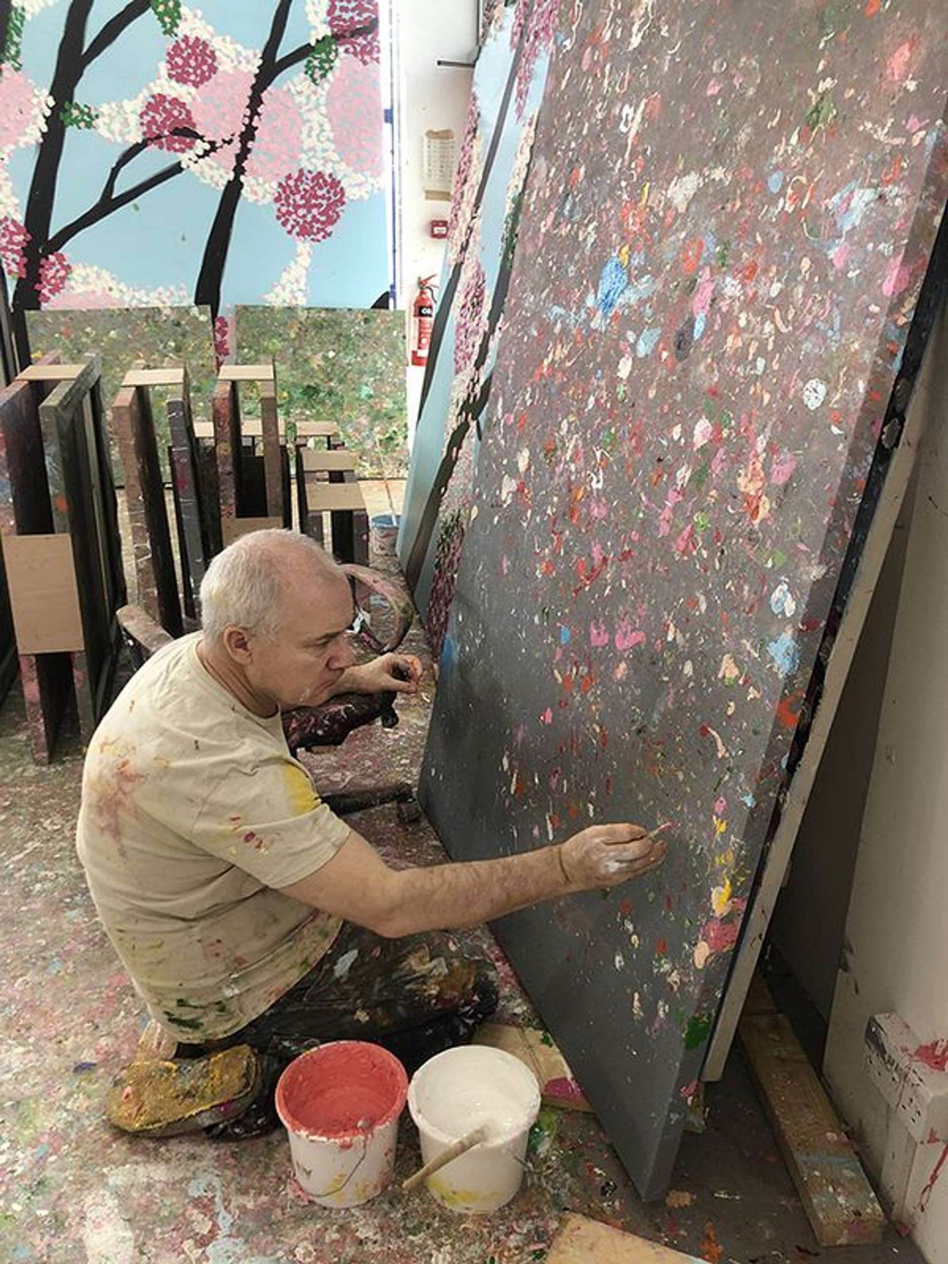 A photograph of the artist Damien Hirst at work in one of his paintings for Where The Land Meets The Sea. He is shown kneeling on the floor, surrounded and splattered by paint, adding splashes to a canvas with his right hand.