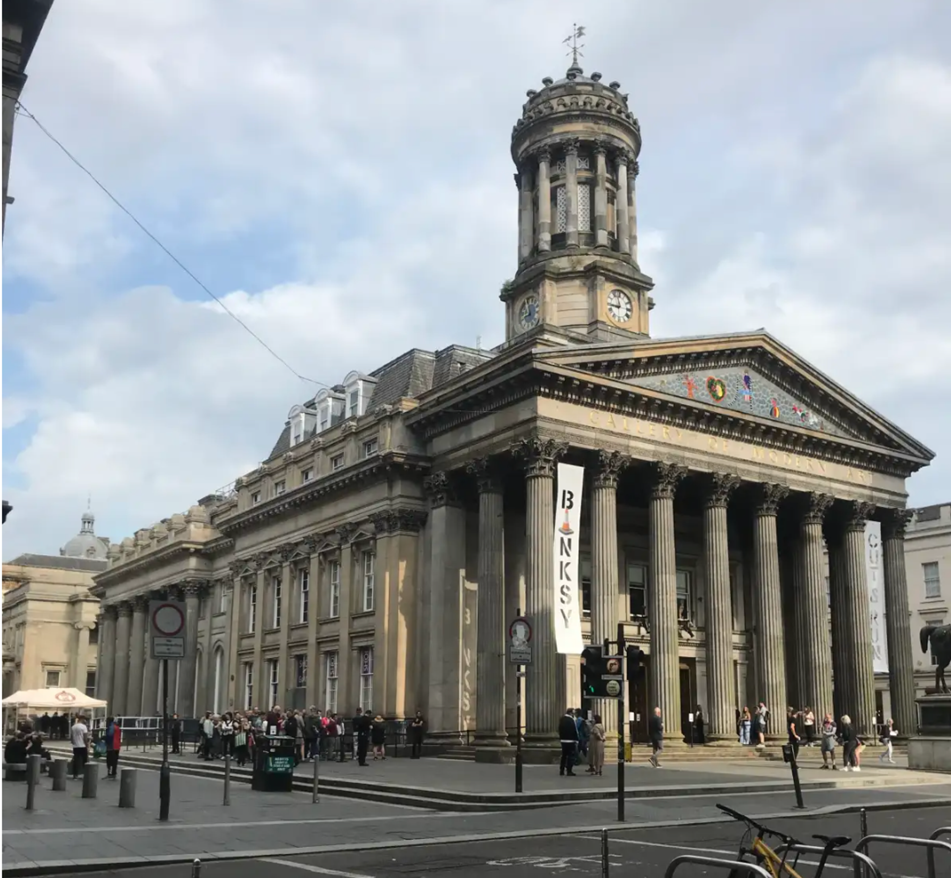 A photo of the outside street entrance to GoMA featuring Banksy's Cut n Run exhibition, displaying a white hanging banner that reads 'Cut N Run' .