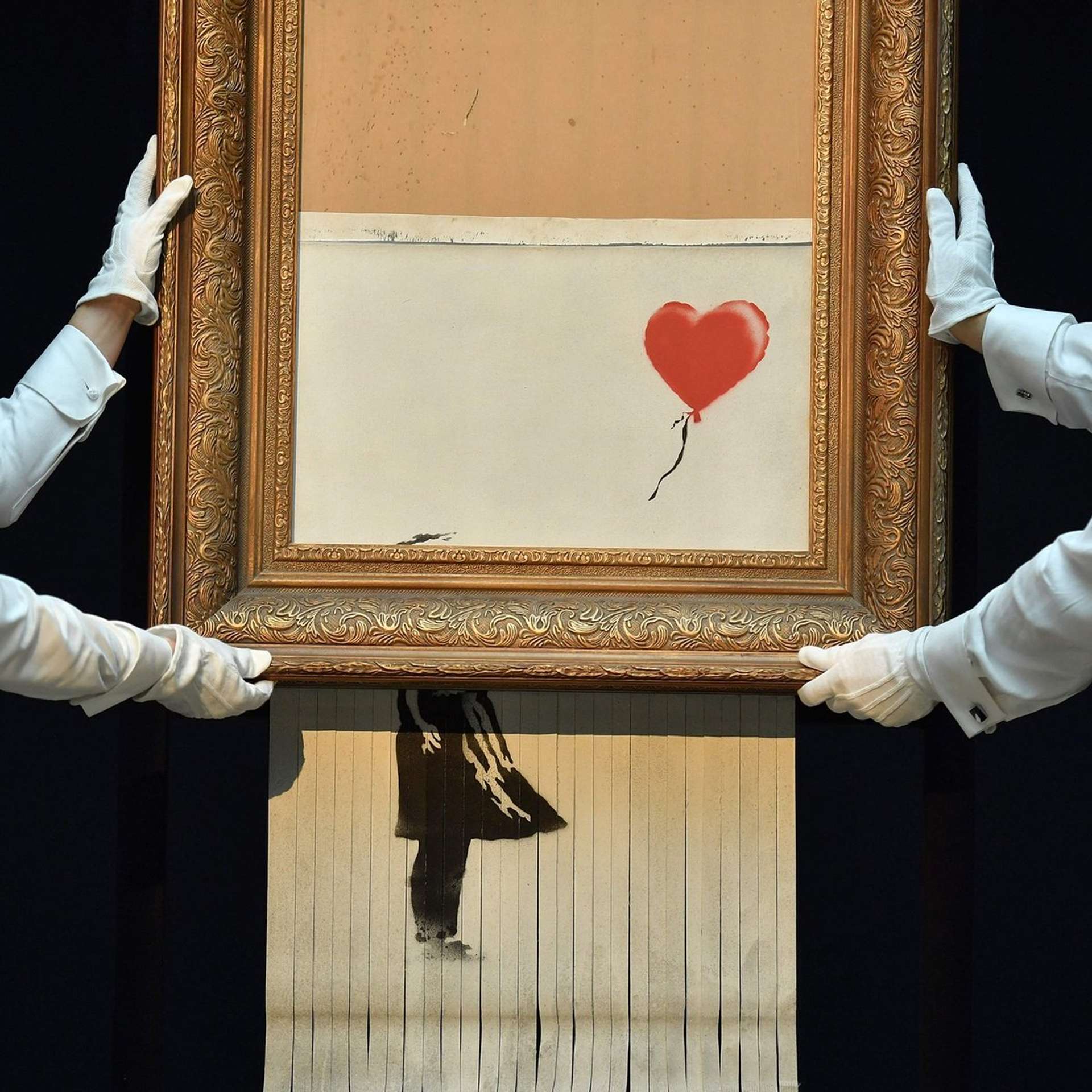 A photograph of two Sotheby's auction staff handling Banksy's shredded Girl With Balloon artwork, which suspends below the gilt frame in shreds.