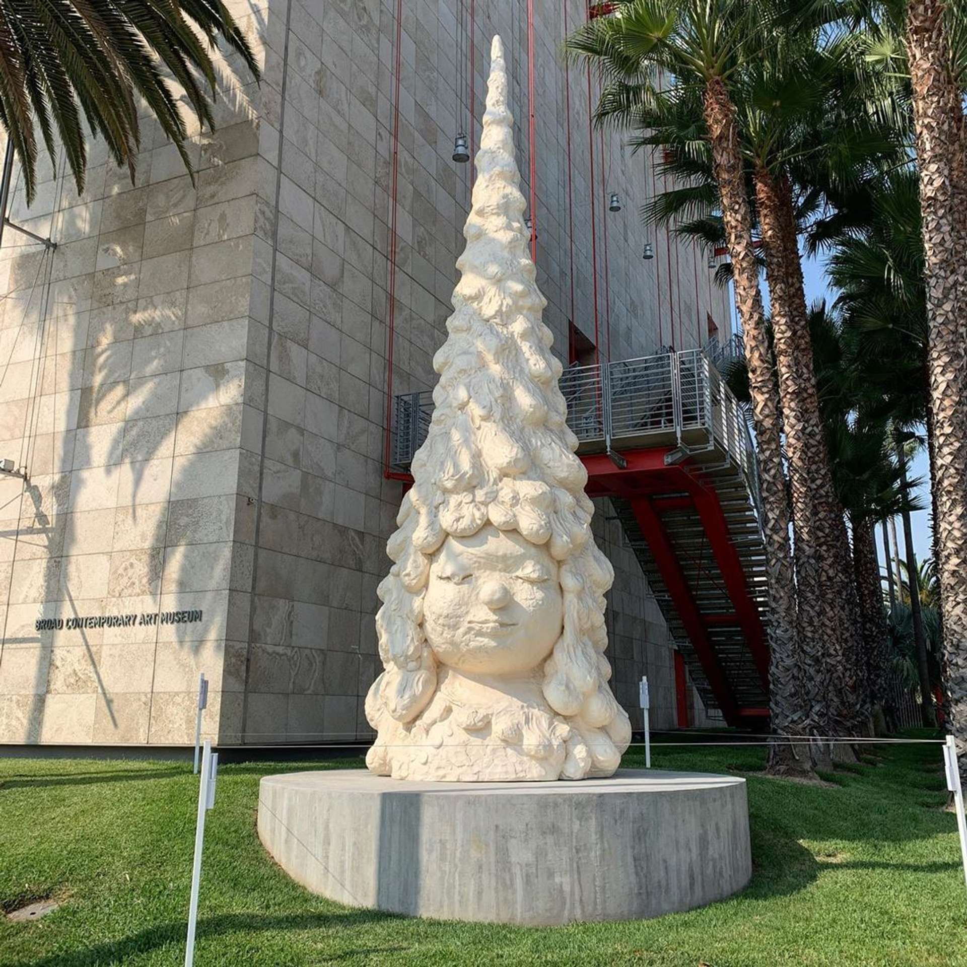 White miss forest sculpture pictured in installation at Los Angeles contemporary art museum. She is shown as a face at the base of a tall tree reminiscent of a pine.
