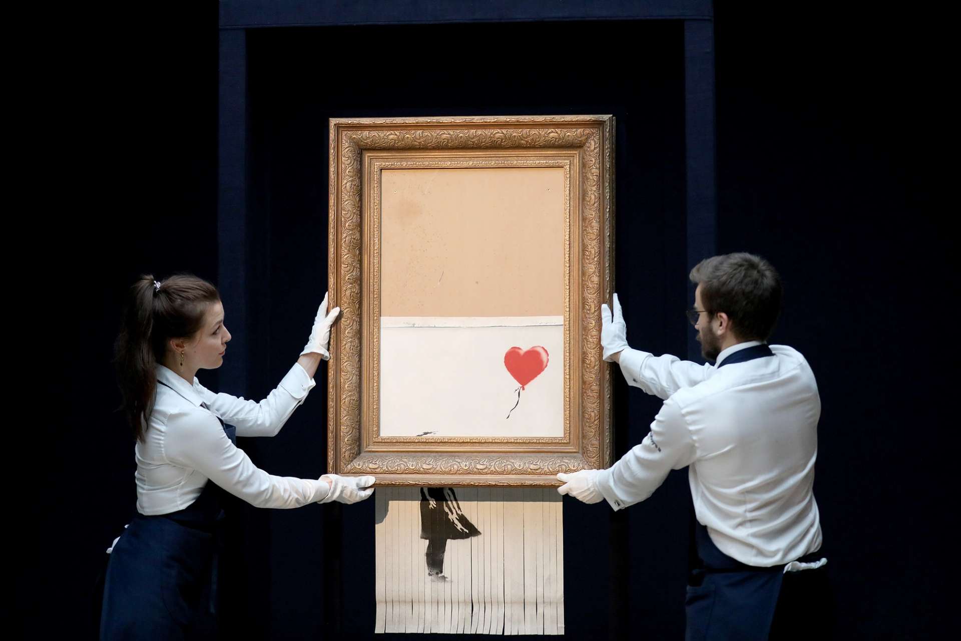 A photograph of two Sotheby's auction staff handling Banksy's shredded Girl With Balloon artwork, which suspends below the gilt frame in shreds.