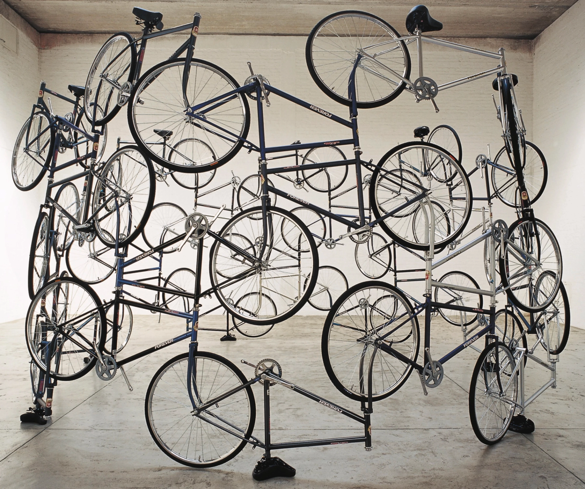 A photograph capturing an intricate sculptural assembly of bicycles, creatively interwoven and locked together through their wheels and handlebars, forming a large-scale sculpture. The artwork is displayed in an empty gallery space.