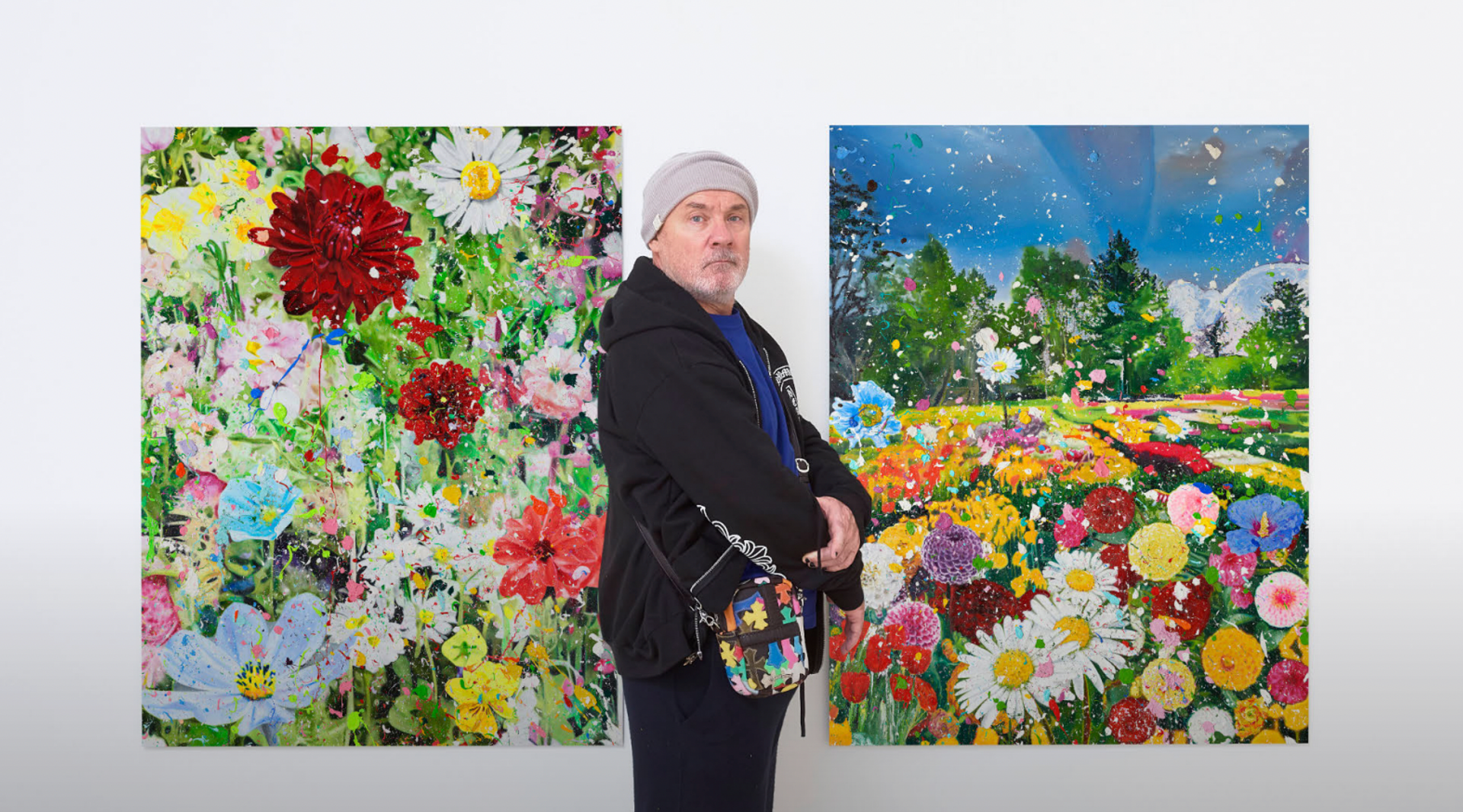 A photograph of Damien Hirst in a tracksuit standing in front of two of his The Secrets prints, which depict a colourful compositions of flowers in a field.
