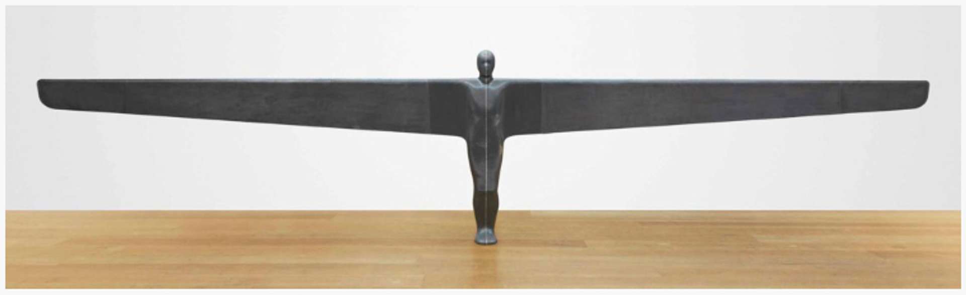 A life-sized standing sculpture of a figure flanked by an 8.5-metre wingspan. The figure has no defining features other than the human form and stands on the ground with its knees slightly bent. The sculpture is captured in a photograph within an empty gallery space, emphasising its presence and allowing viewers to appreciate its form and proportions.