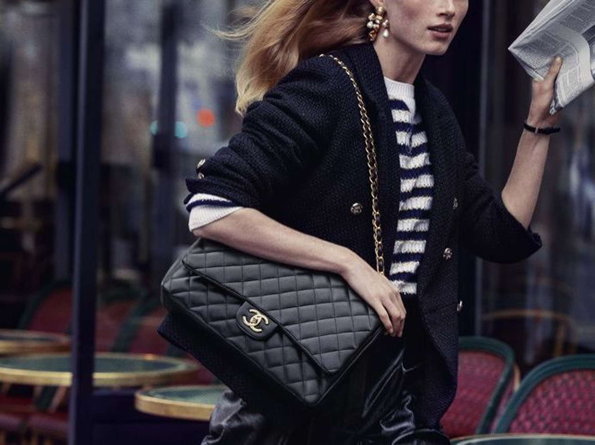 An image of a model, dressed in a striped top and a cardigan, holding a large black Chanel Classic Flap Bag.