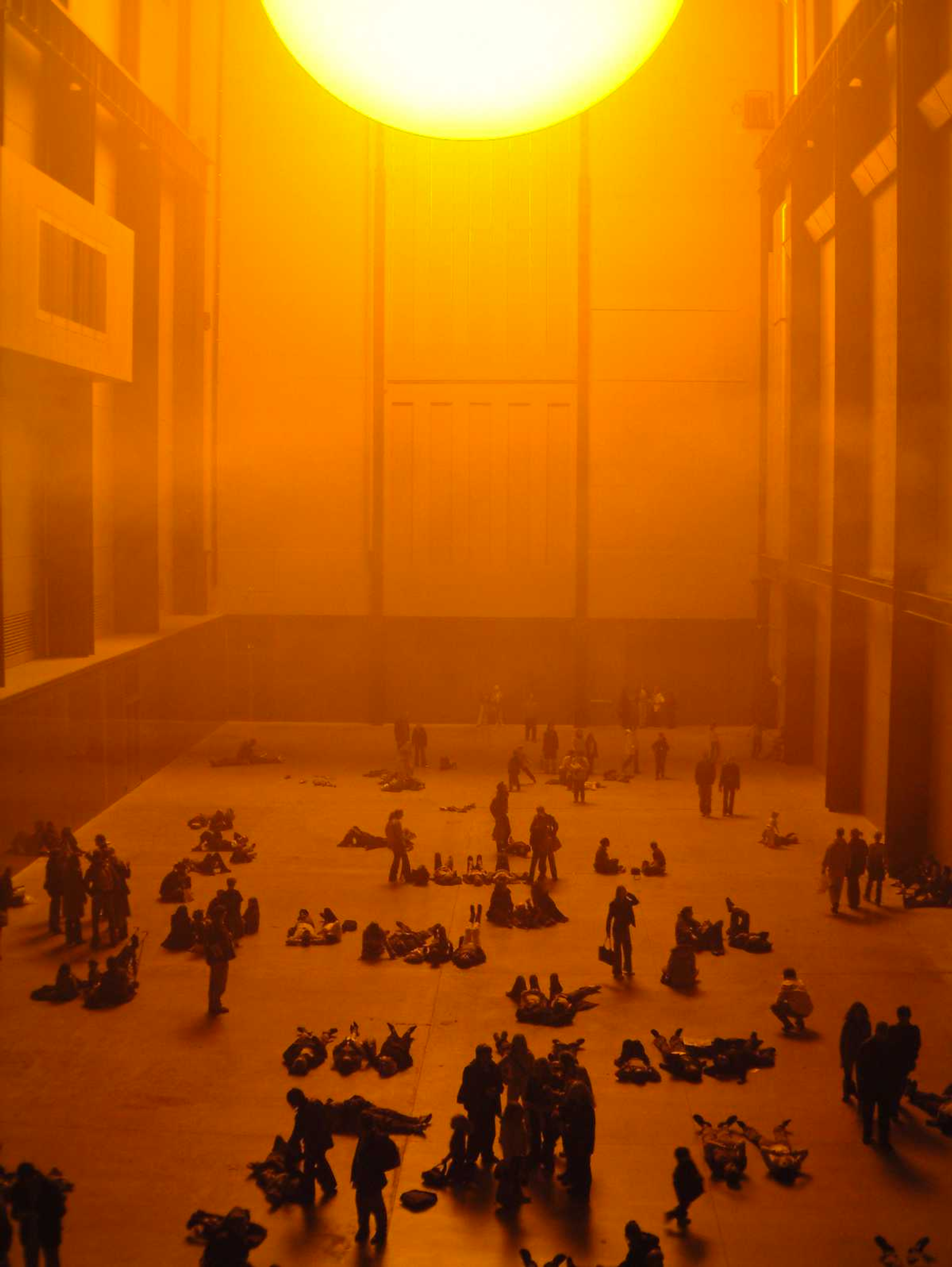 This photograph shows the Tate's Turbine Hall, a large room bathed in a yellow glowing light emanating from a large orb in the ceiling. Several people can be seen standing, sitting or lying down beneath it.