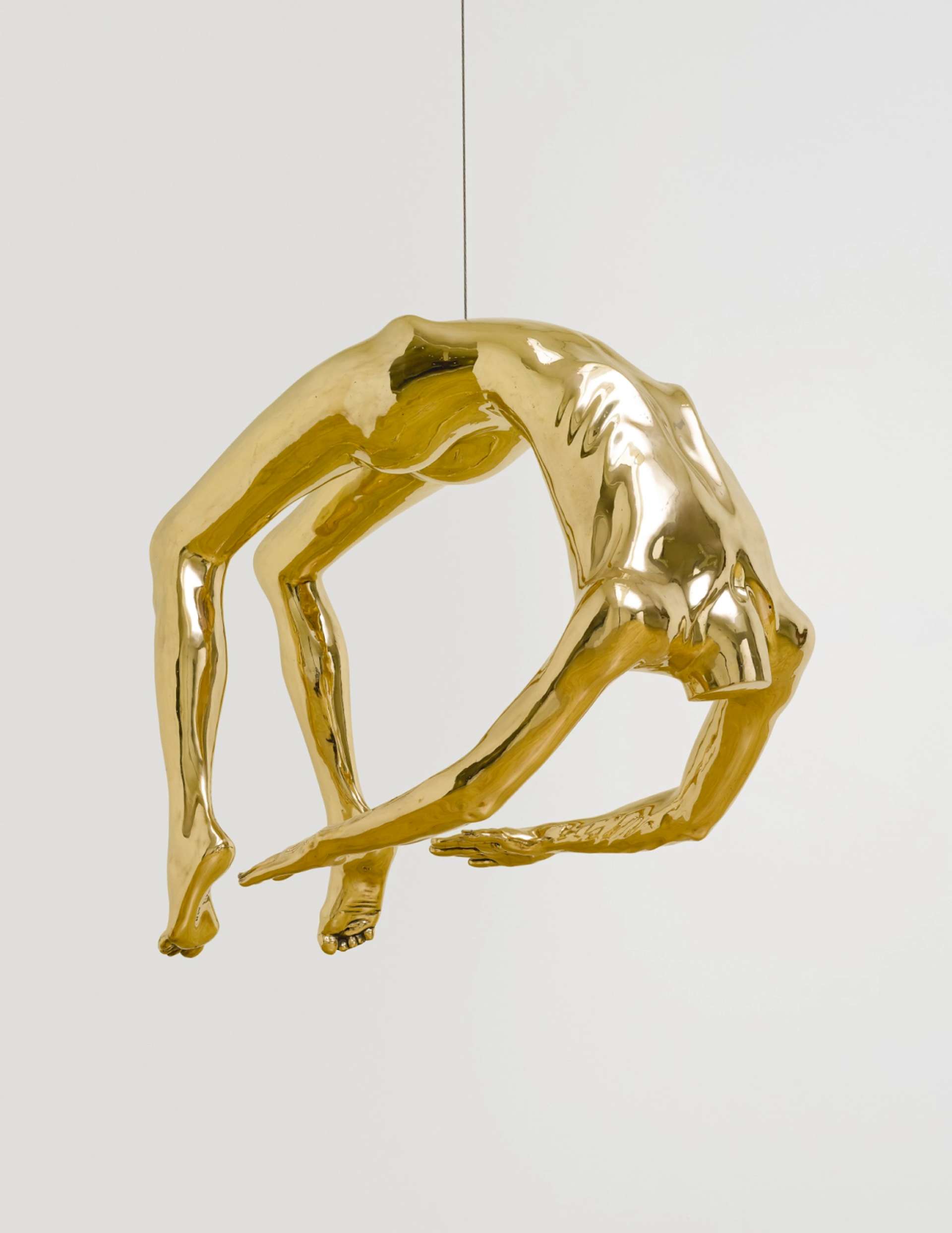 A polished bronze sculpture suspended from a barely visible string, depicting a headless body in an exaggerated arched pose. Its arms are extended and reaching towards its feet, creating a geometric circle shape.