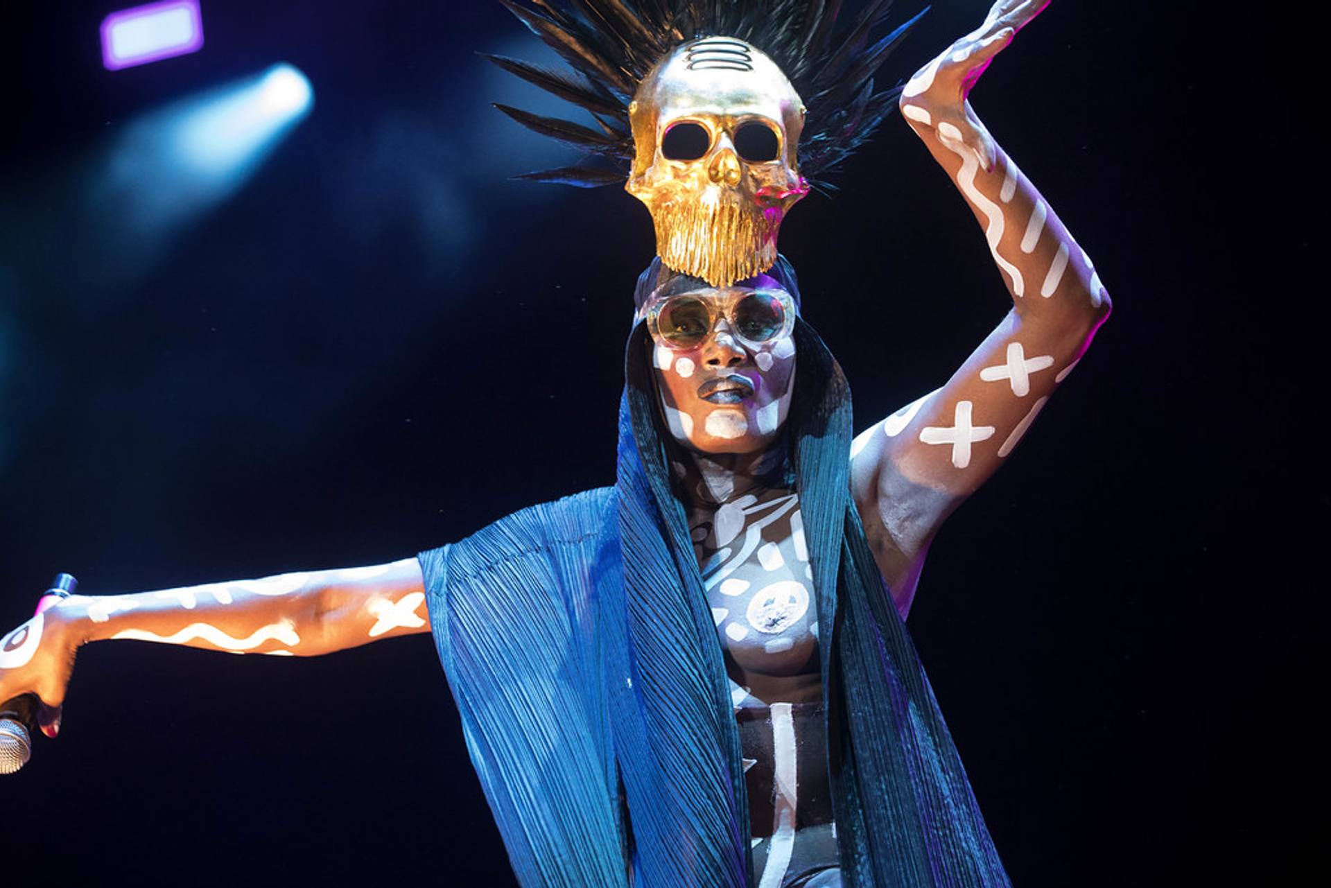 An image of performer Grace Jones on stage, wearing a golden skull atop her head. He is wearing sunglasses and glaring at the camera.