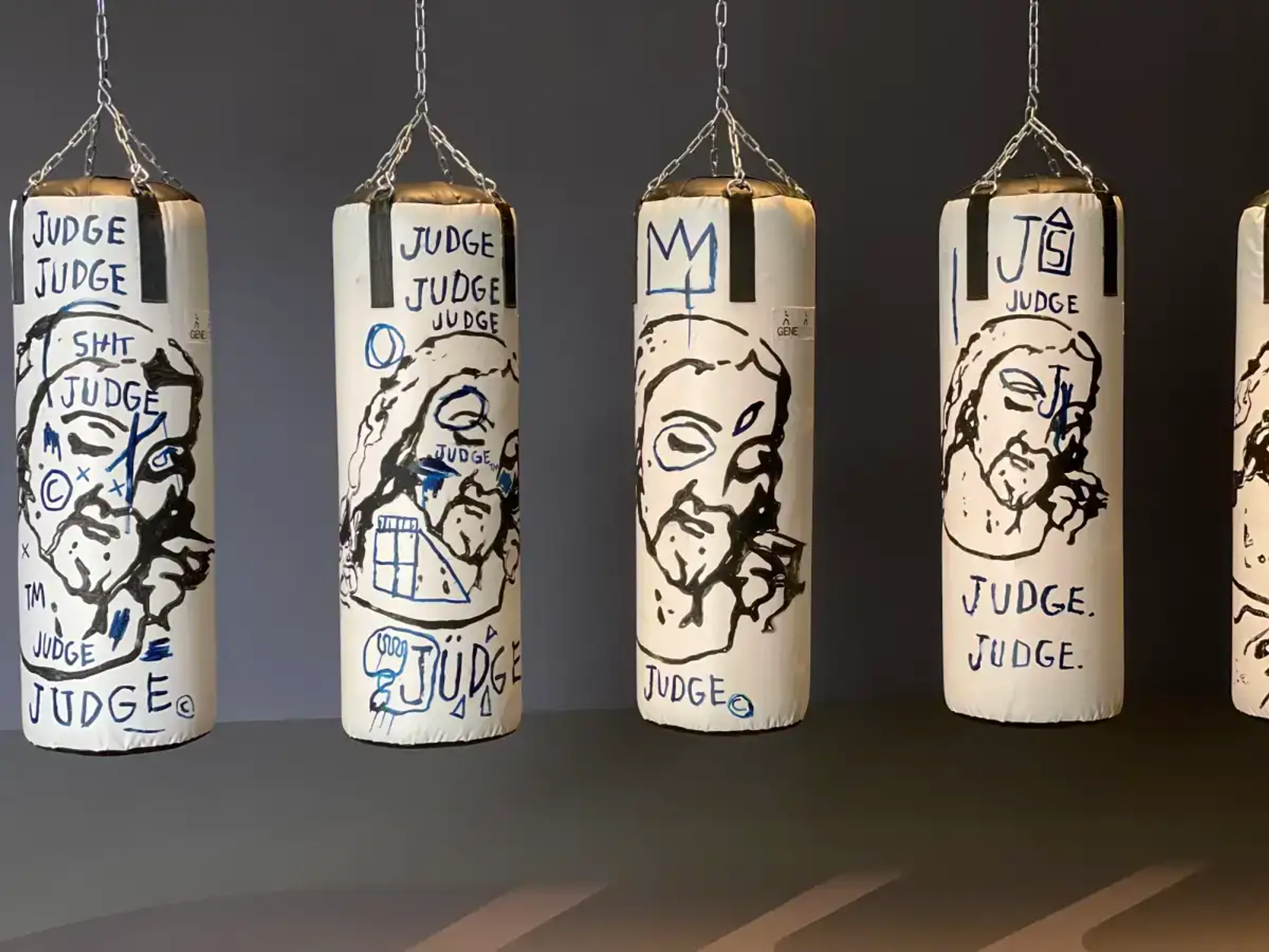 This photograph shows four of the twn punching bags from Warhol and Basquiat's work. Each is painted with a depiction of Jesus, which has been variously altered with the word Judge.