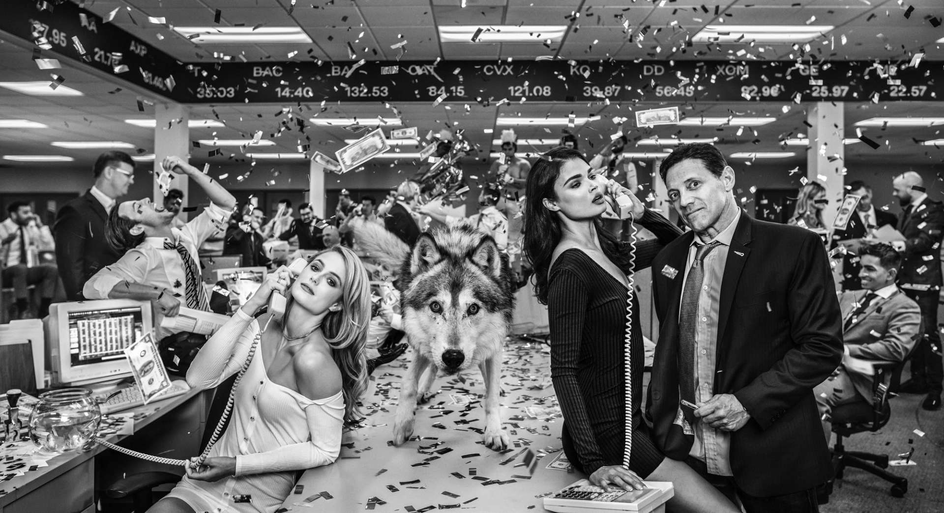 A black and white photograph by artist David Yarrow, showing the “Wolf of Wall Street” Jordan Belfort standing alongside models and an actual wolf, while confetti and money falls from the sky.