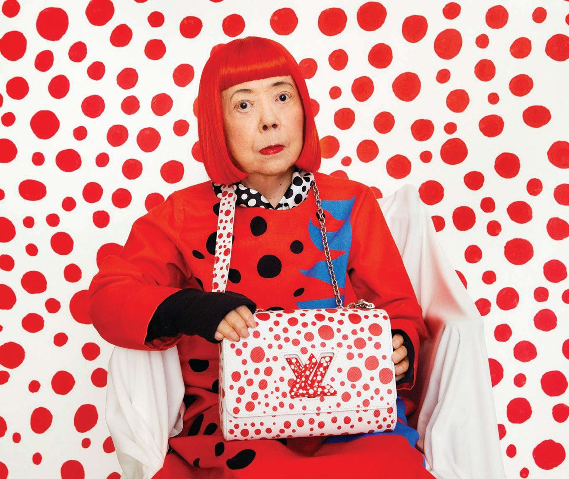 An image of artist Yayoi Kusama, wearing red, holding one of the bags she designed in collaboration with Louis Vuitton.