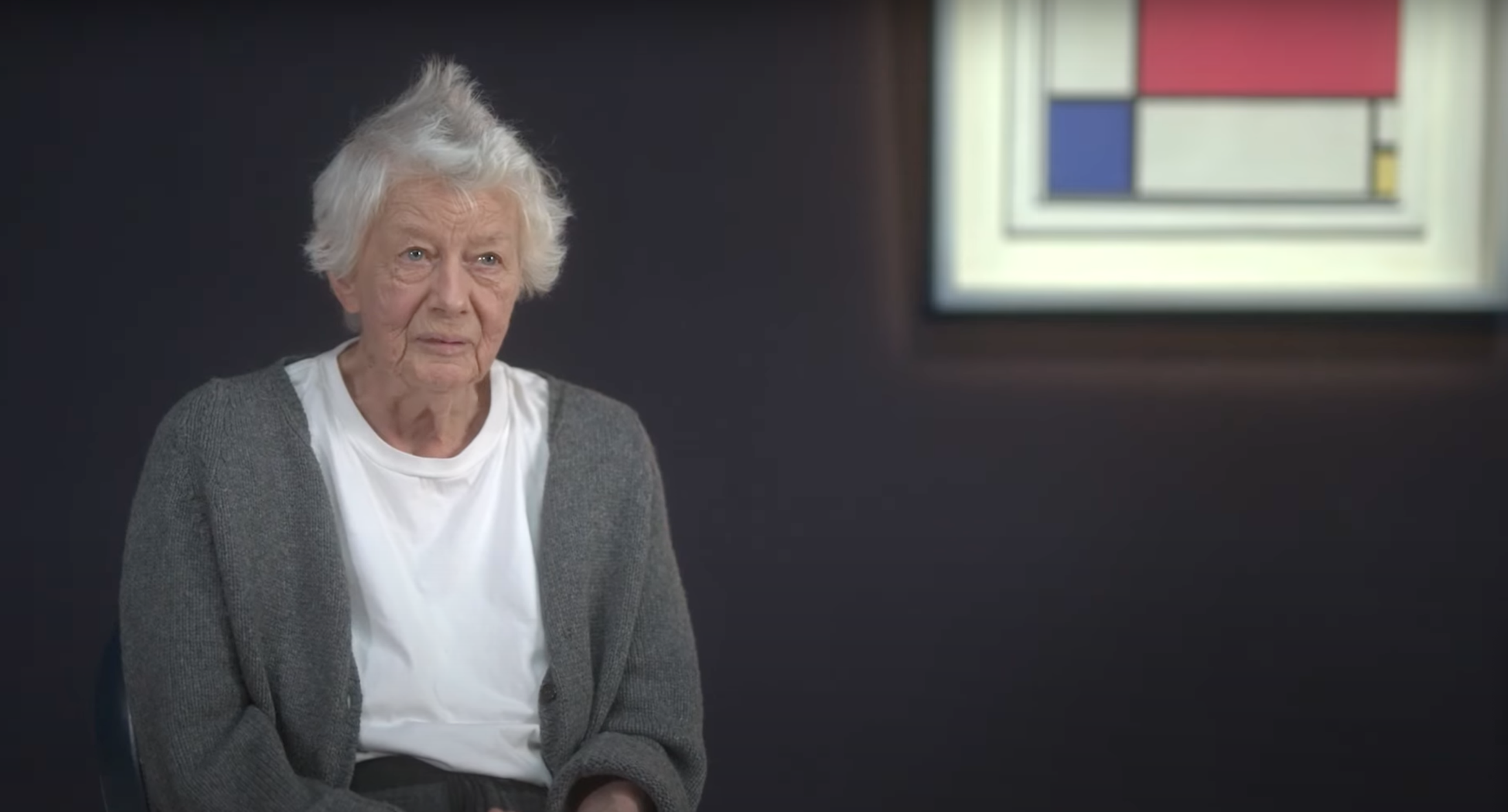 This screenshot shows artist Bridget Riley sitting in front of the work Composition No. II by Piet Mondrian.