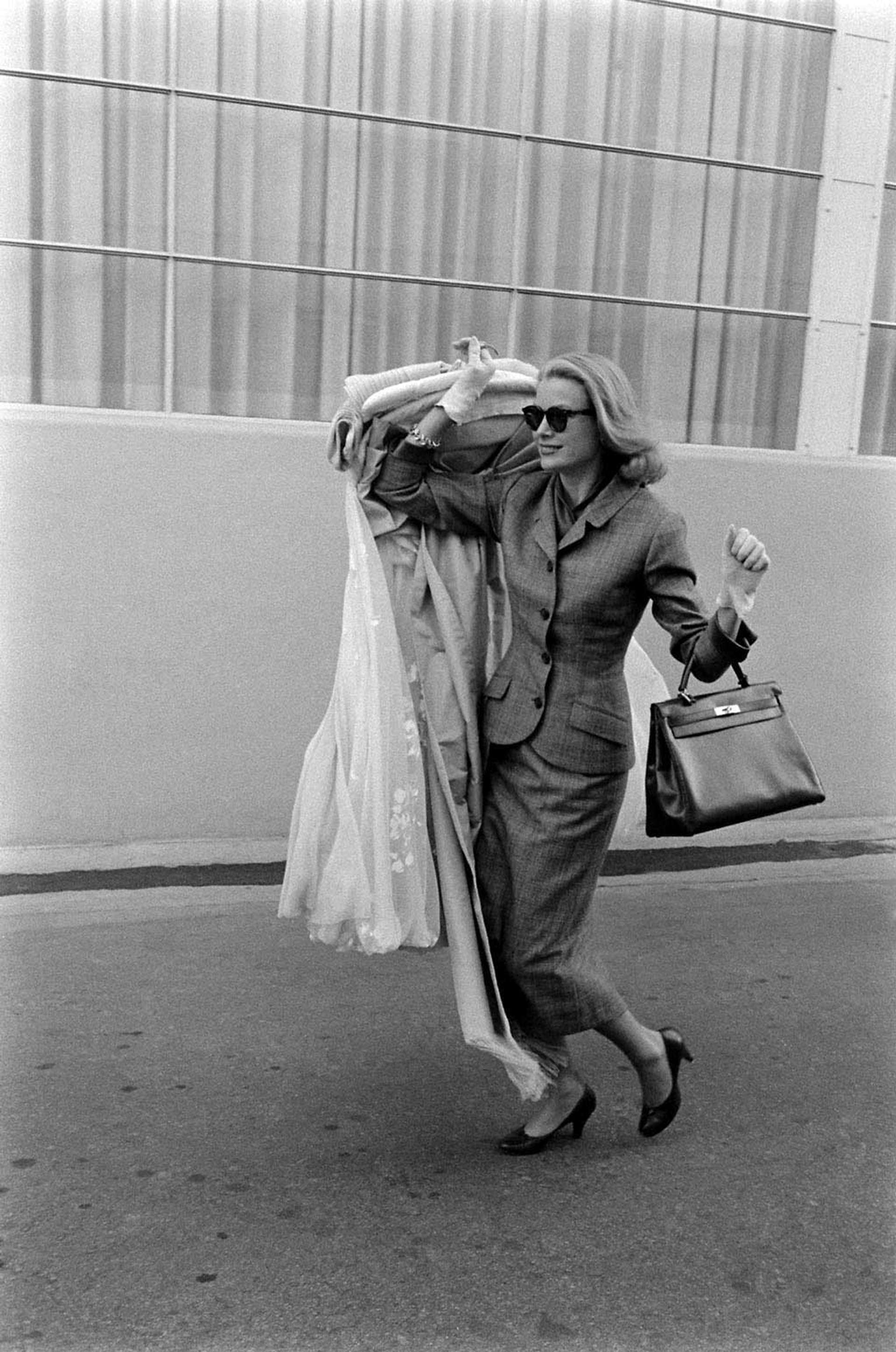 An image of actress Grace Kelly, carrying several clothes and a Kelly bag, leaving the Hollywood Studios she worked on for the last time.]