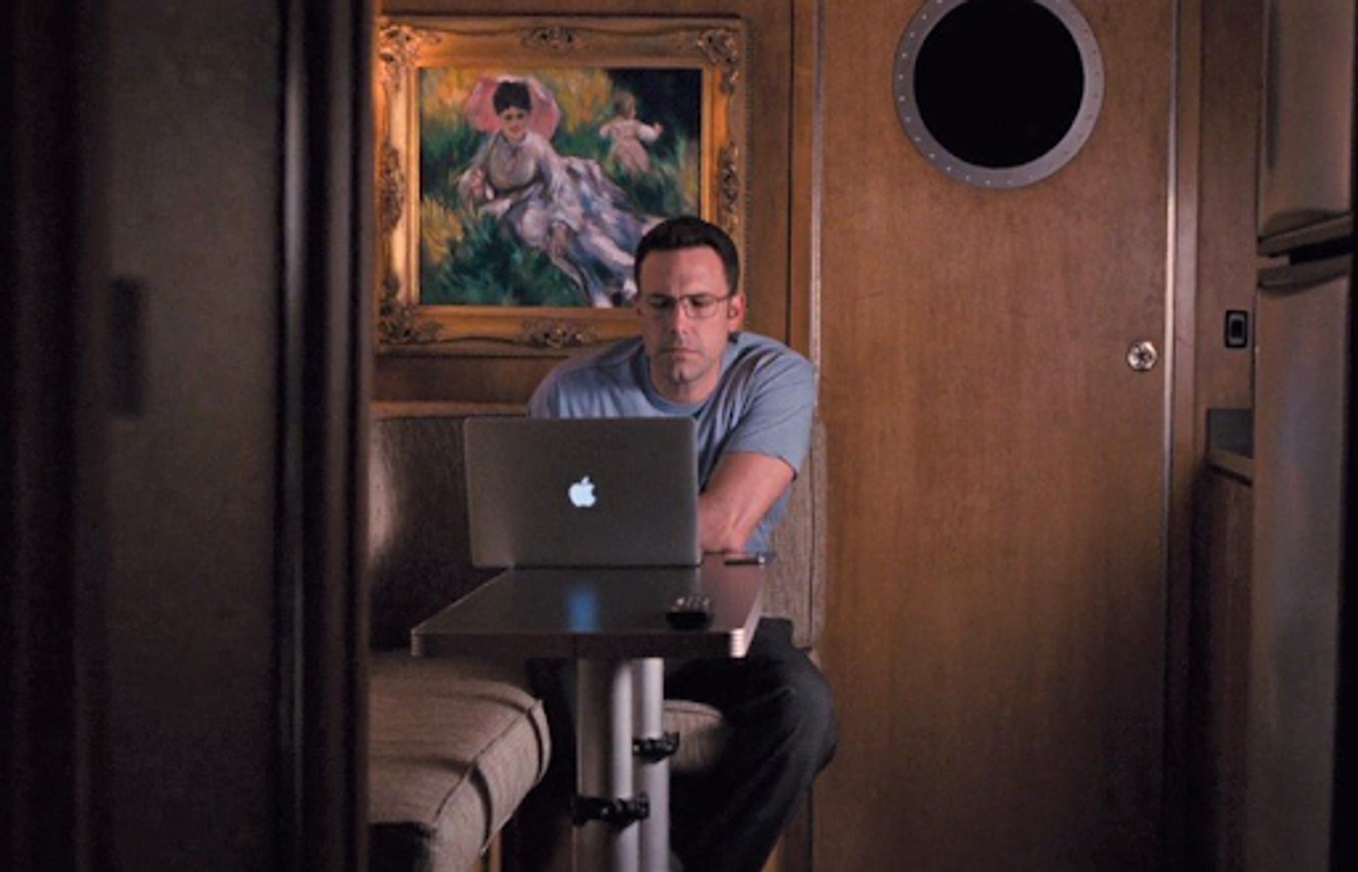 Photograph from the film The Accountant. Man sits at a table with his laptop open. There is a fine art painting behind him. 