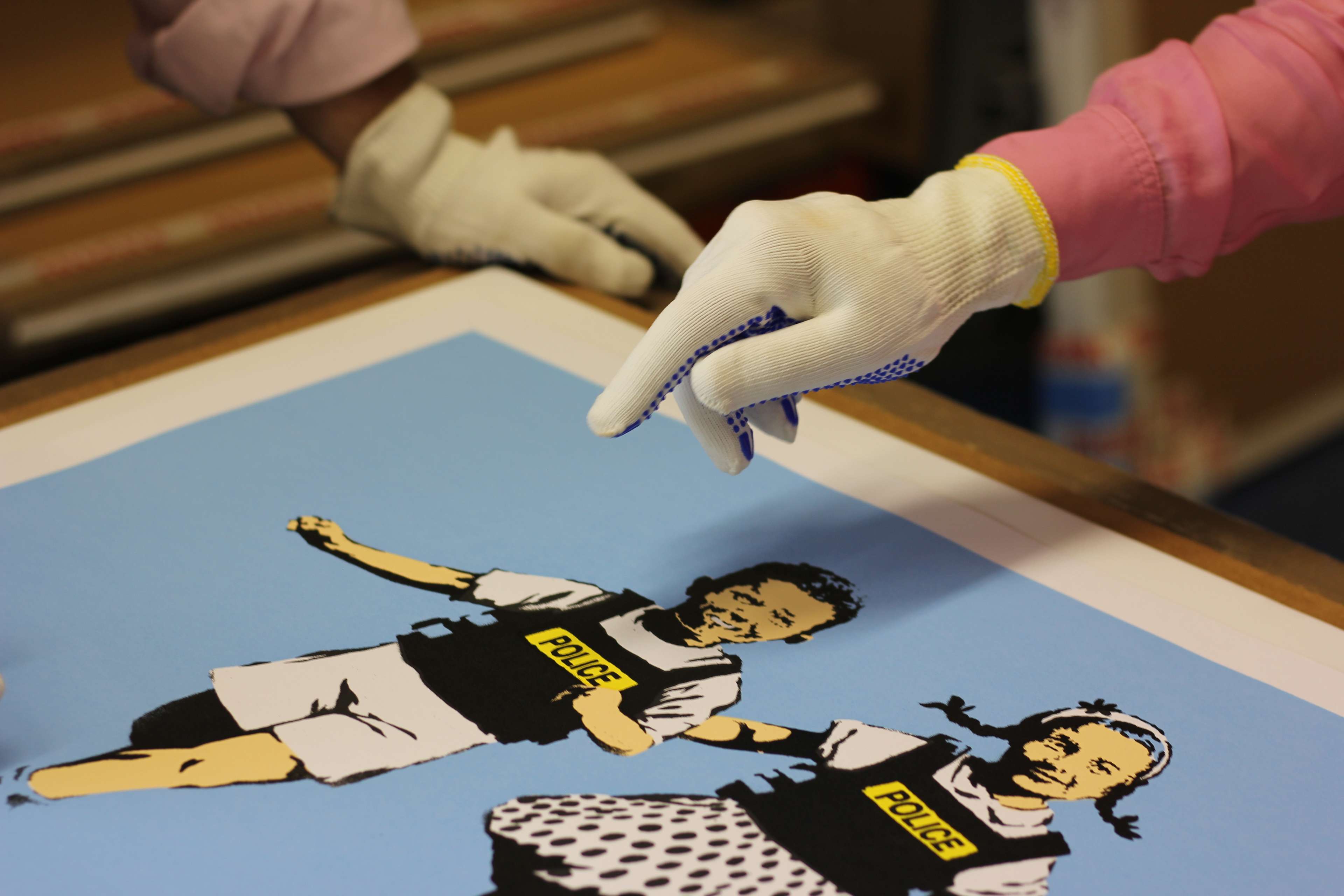 Two people wearing white gloves, with one reaching and pointing towards a paper artwork depicting a young girl and boy wearing police vests against a sky blue background.