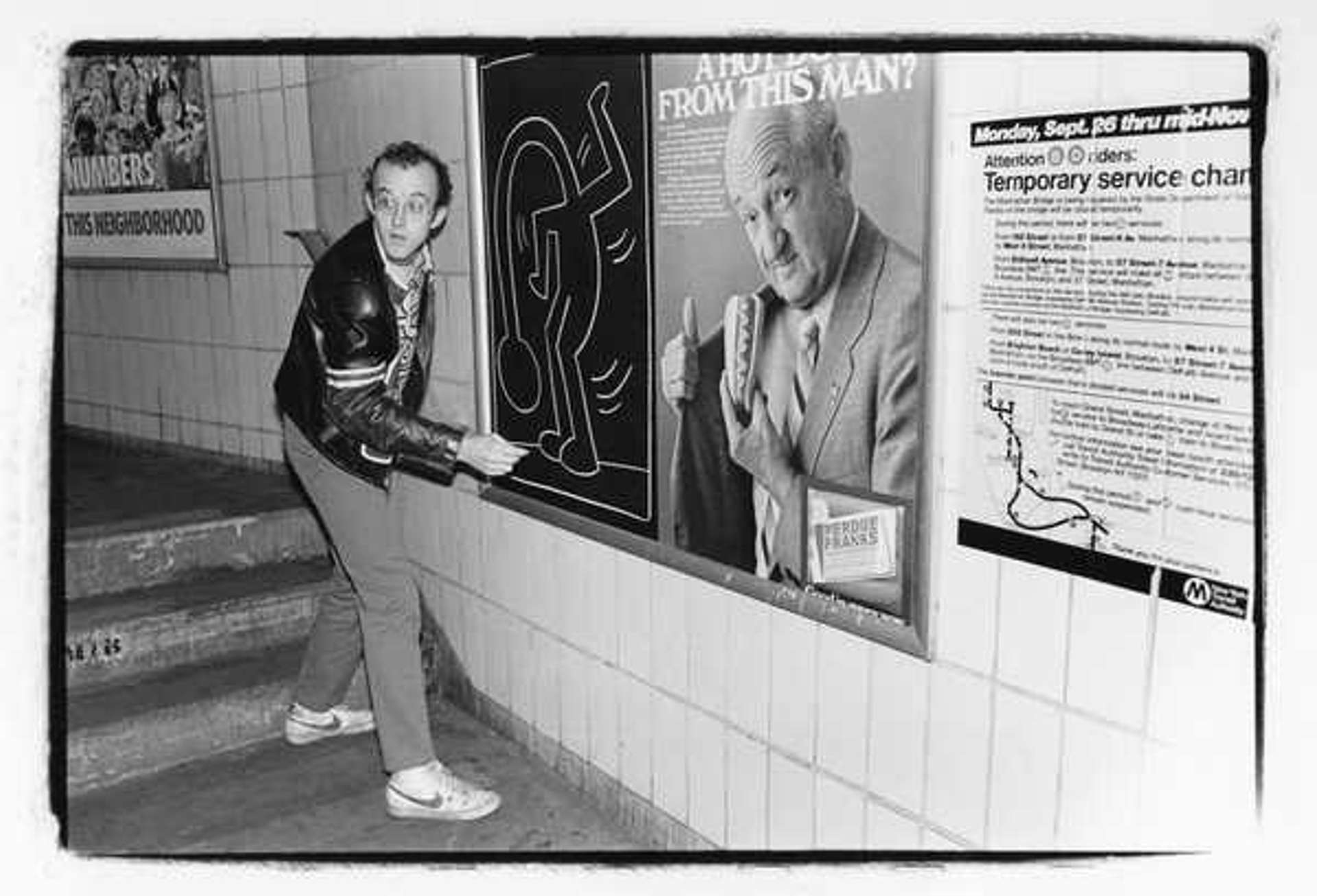 Keith Haring at work on one of his Subway Drawings in New York. The artist poses with his chalk by the black and white drawing, which depicts a dancing figure with a long drooping neck.