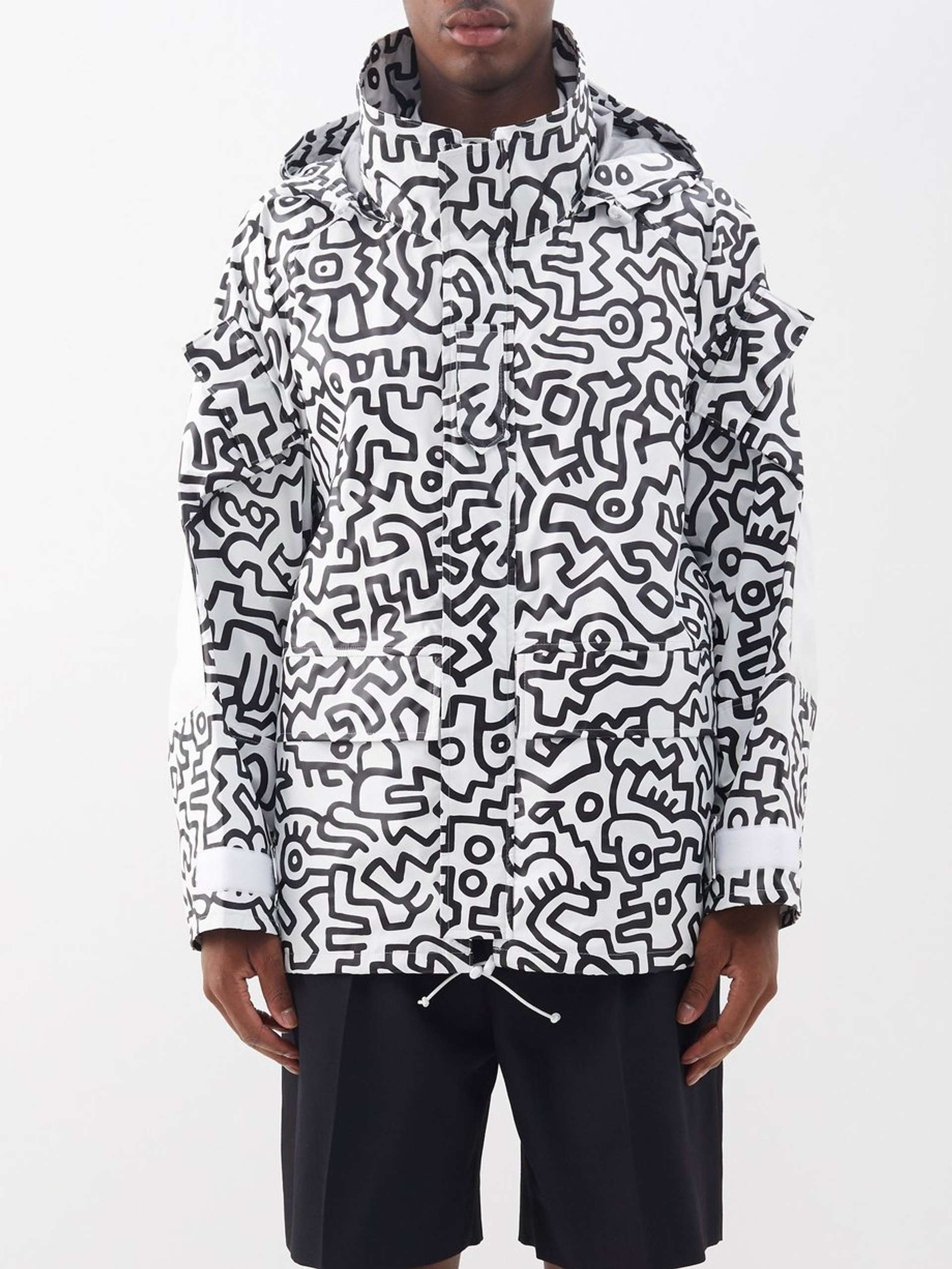 An image of a model wearing a white windbreaker Junya Watanabe jacket that is completely covered in Keith Haring’s signature doodles in black.