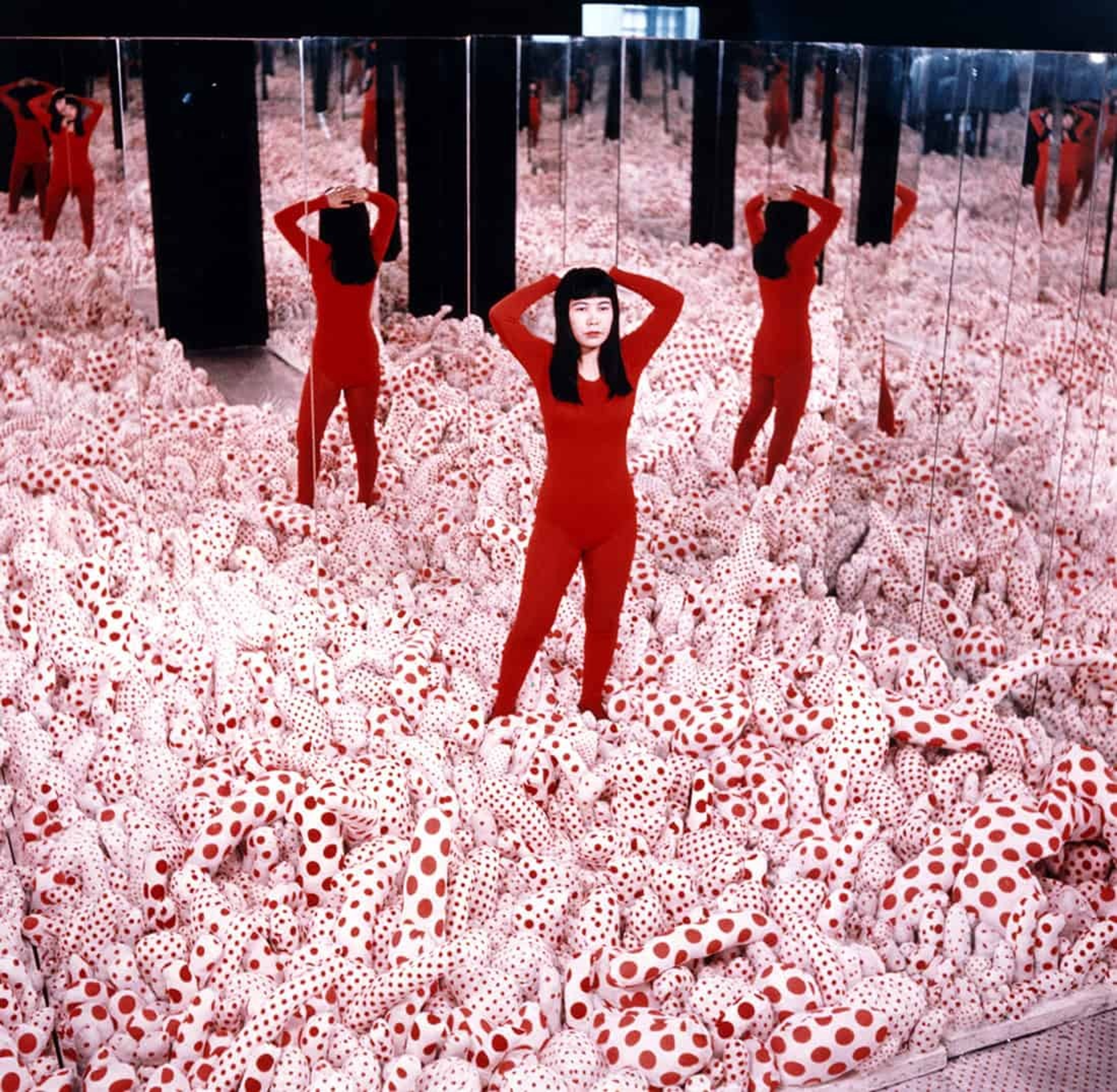 Yayoi Kusama standing inside an Infinity Mirror Room filled with red and white polka-dotted forms.