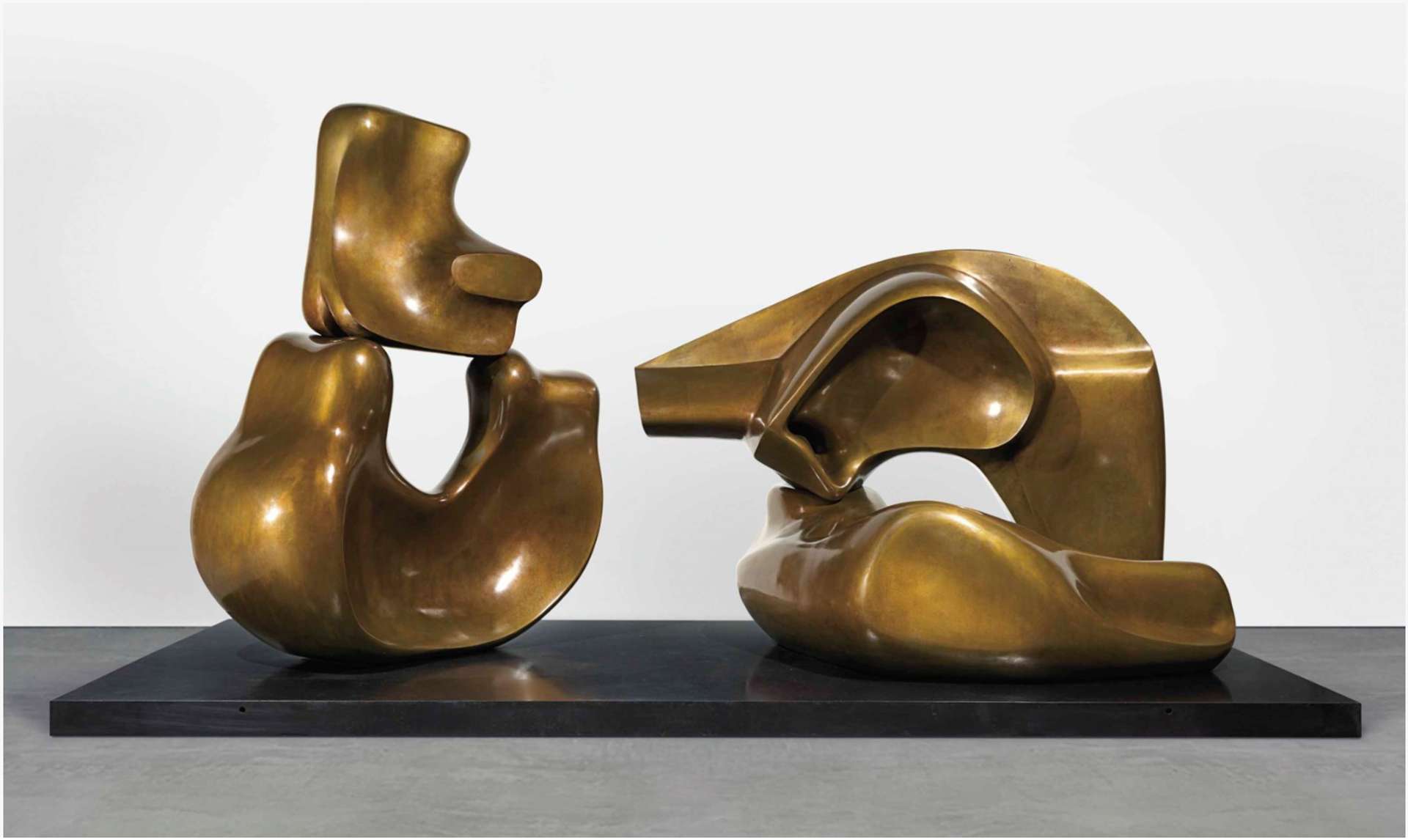 Henry Moore's bronze sculpture featuring four fragments arranged in pairs. On the left side, two pieces are placed together, with a smaller fragment balancing on the edges of a larger bottom piece. The larger piece showcases a concaved void, adding depth and balance to the composition. On the right side, there are two additional pieces: one laid down in an L-shape, while the other extends upward and crosses over the first shape at a right angle.