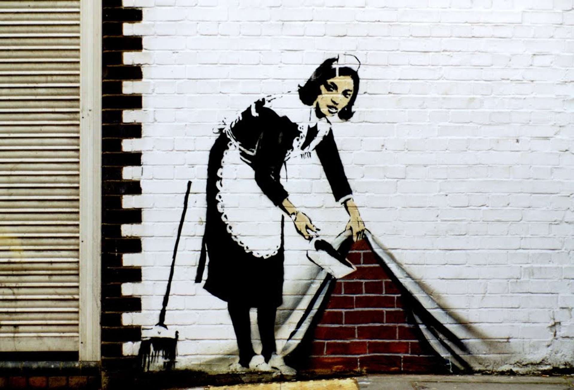 A street art mural by Banksy depicting a woman in a maid uniform, appearing to lift a white curtain to reveal the brick wall underneath where she discards her sweepings.