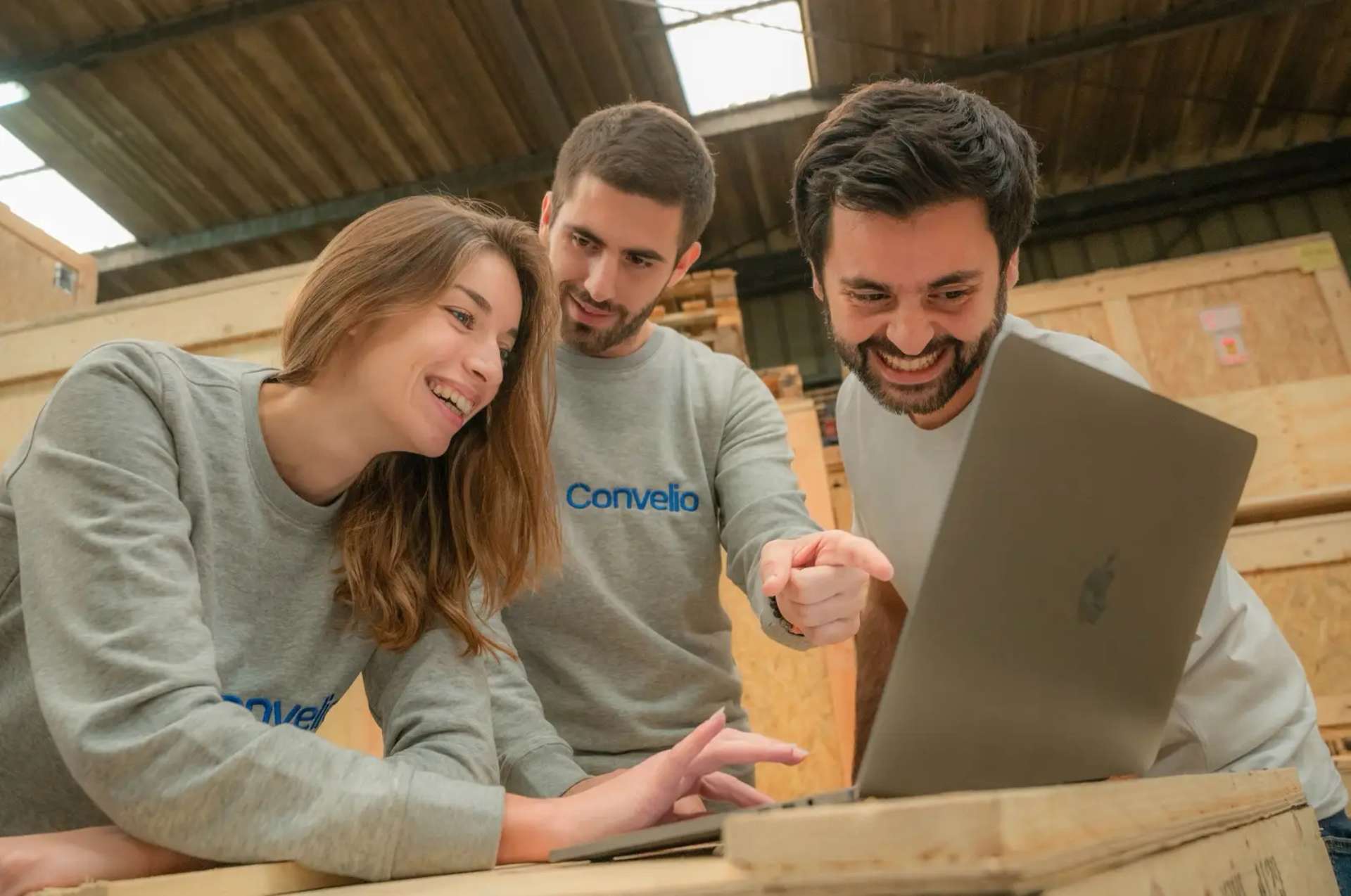 Three members of the Convelio team in a warehouse looking at a computer screen, all wearing a grey sweatshirt with the company name and logo in blue.