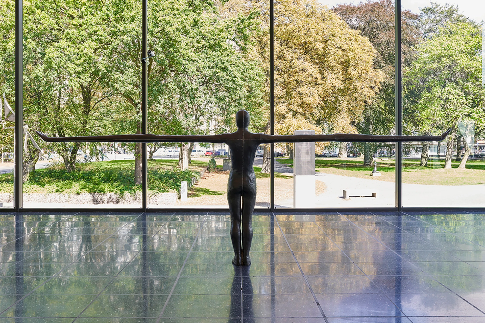 A photograph showing the back view of a sculpture representing the human figure. The figure stands firmly with its feet grounded while being supported by an imposing wingspan. The knees are slightly bent, adding to the sense of balance and stability. The sculpture is captured from behind, highlighting its dynamic form and captured in a pristine gallery space. The gallery features large glass windows that offer a glimpse of the surrounding trees outside, creating a harmonious connection between the artwork and nature.