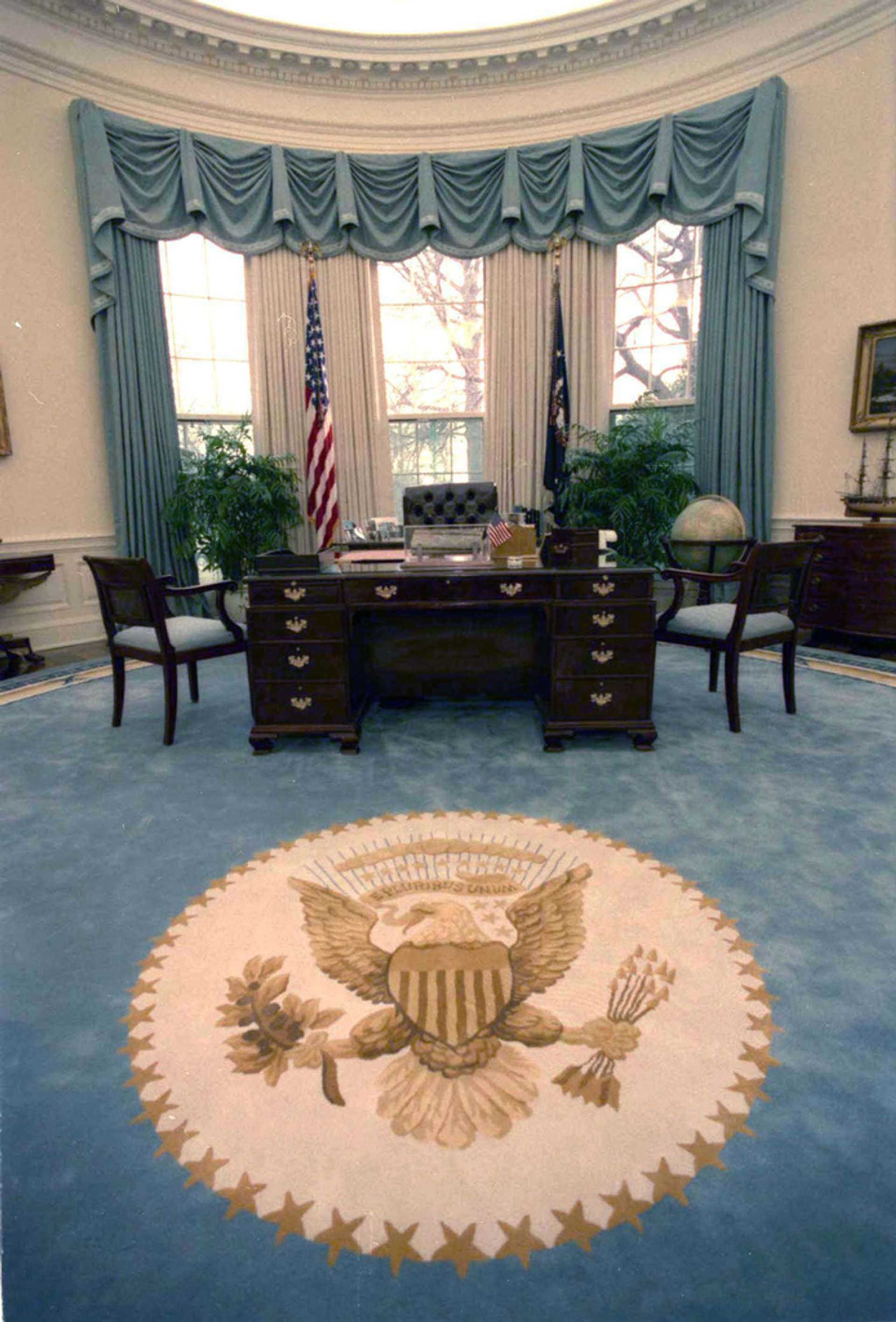 An interior photograph of the oval office during the Bush Administration. A room with blue carpeting and matching blue drapes, with dark furniture.