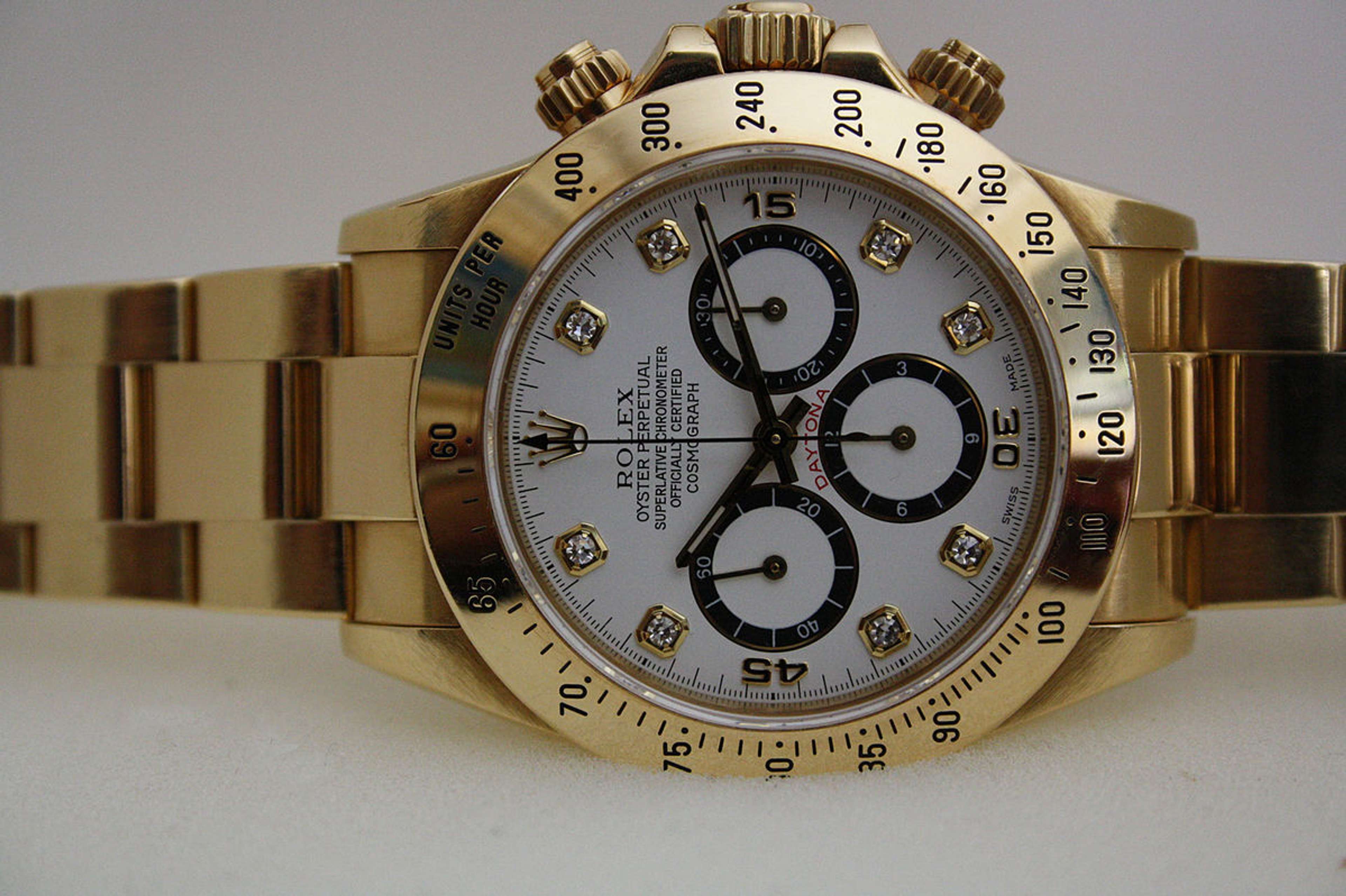 A Rolex Daytona, first launched in 1963. Set against a white background, the watch has a gold metal strap and a white face with three subdials.