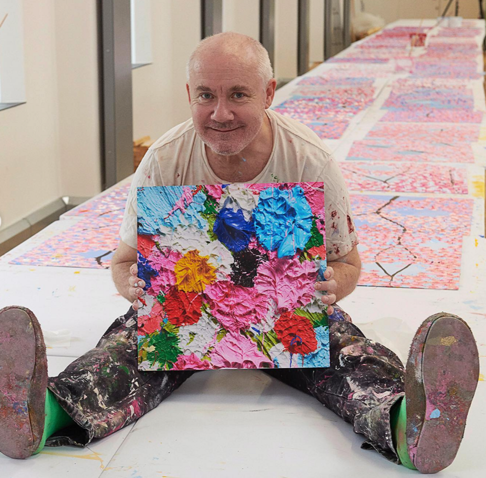 An image of the artist Damien Hirst, holding his work H8-1 Fruitful. In the background, his Cherry Blossom paintings can be seen.