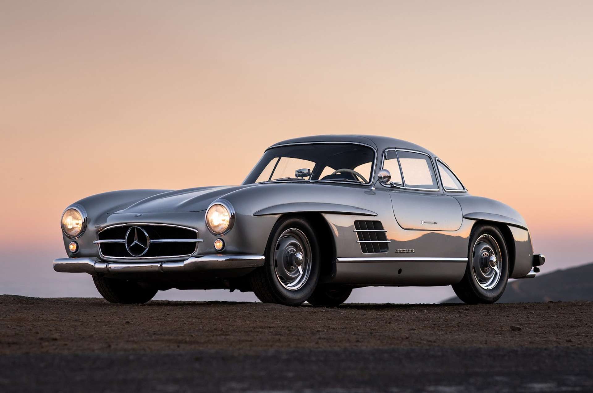1955 silver Mercedes-Benz 300 SL Alloy Gullwing against a sunset background.