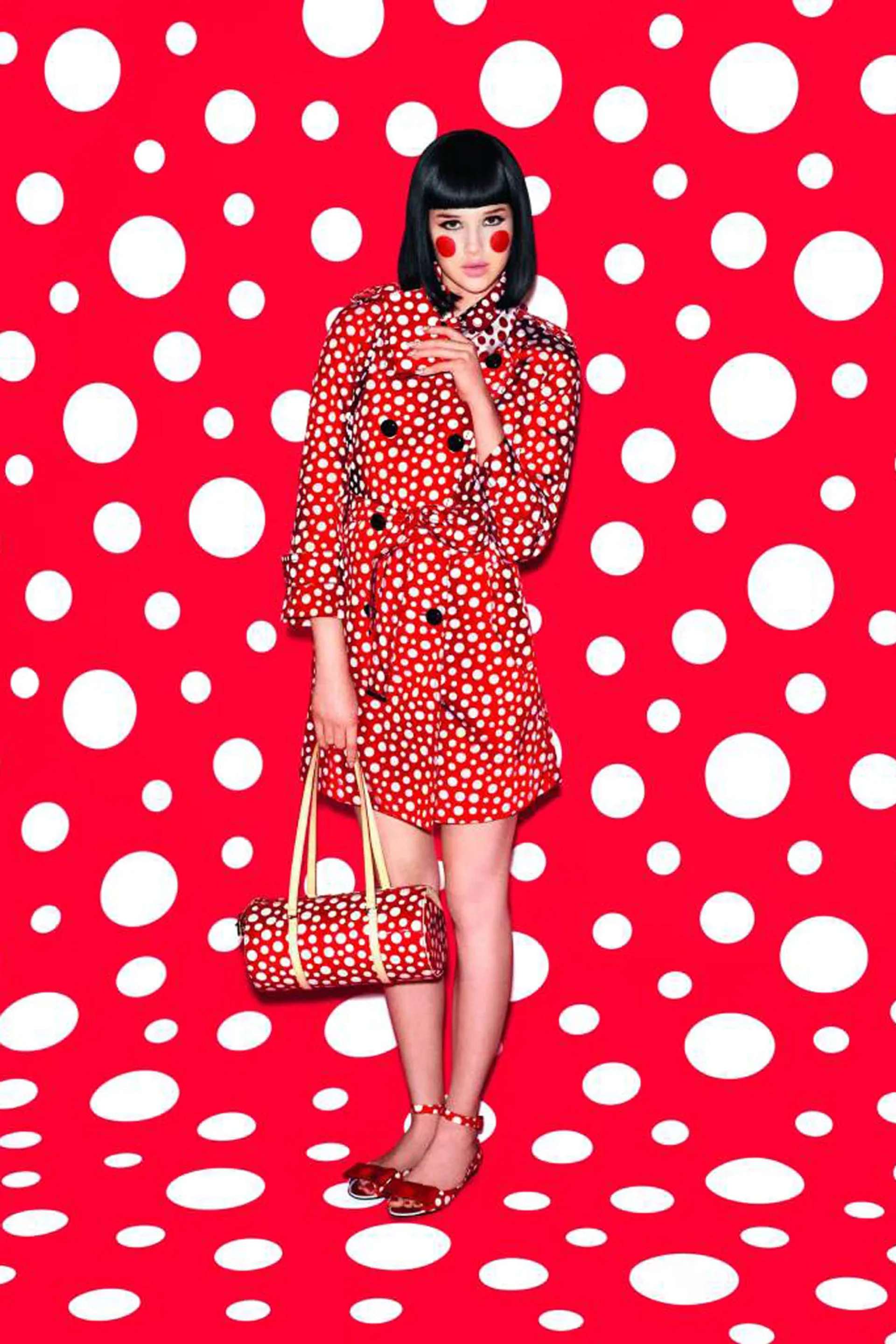 An image of a model dressed in white polka dots on a red background, holding a bag with the same pattern by Yayoi Kusama. She is standing against a background featuring the same print.