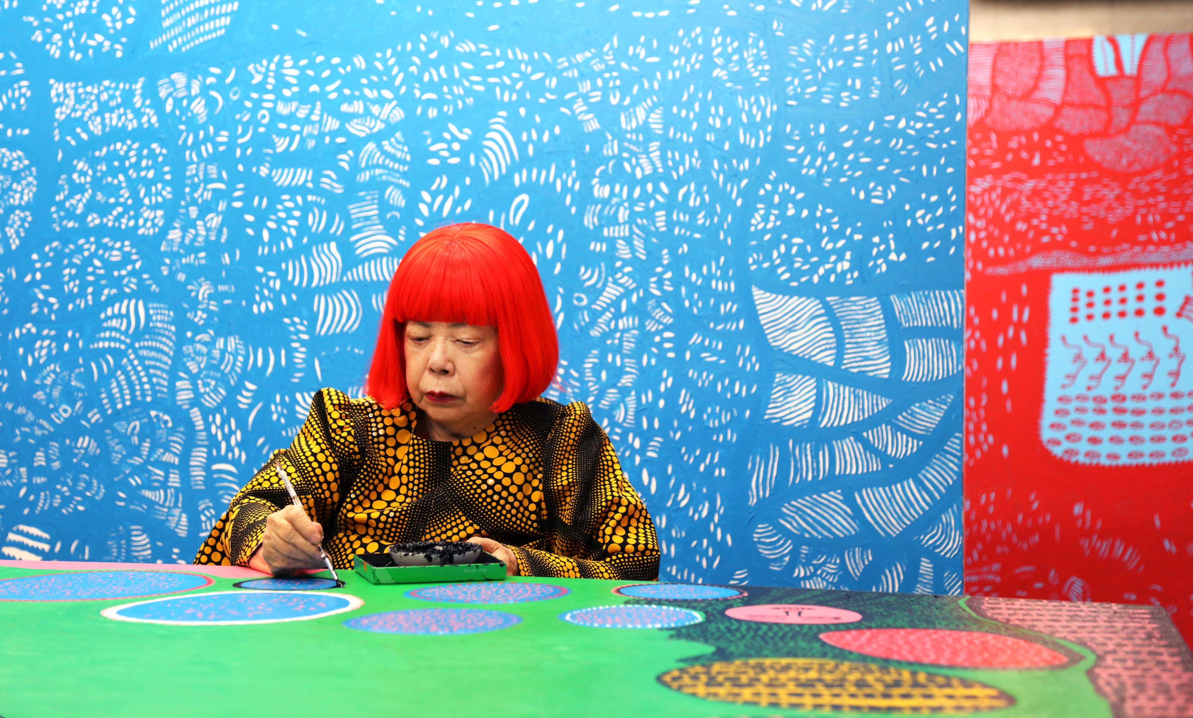 Yayoi Kusama working at a table in her Tokyo studio, surrounded by brightly coloured patterned artworks.
