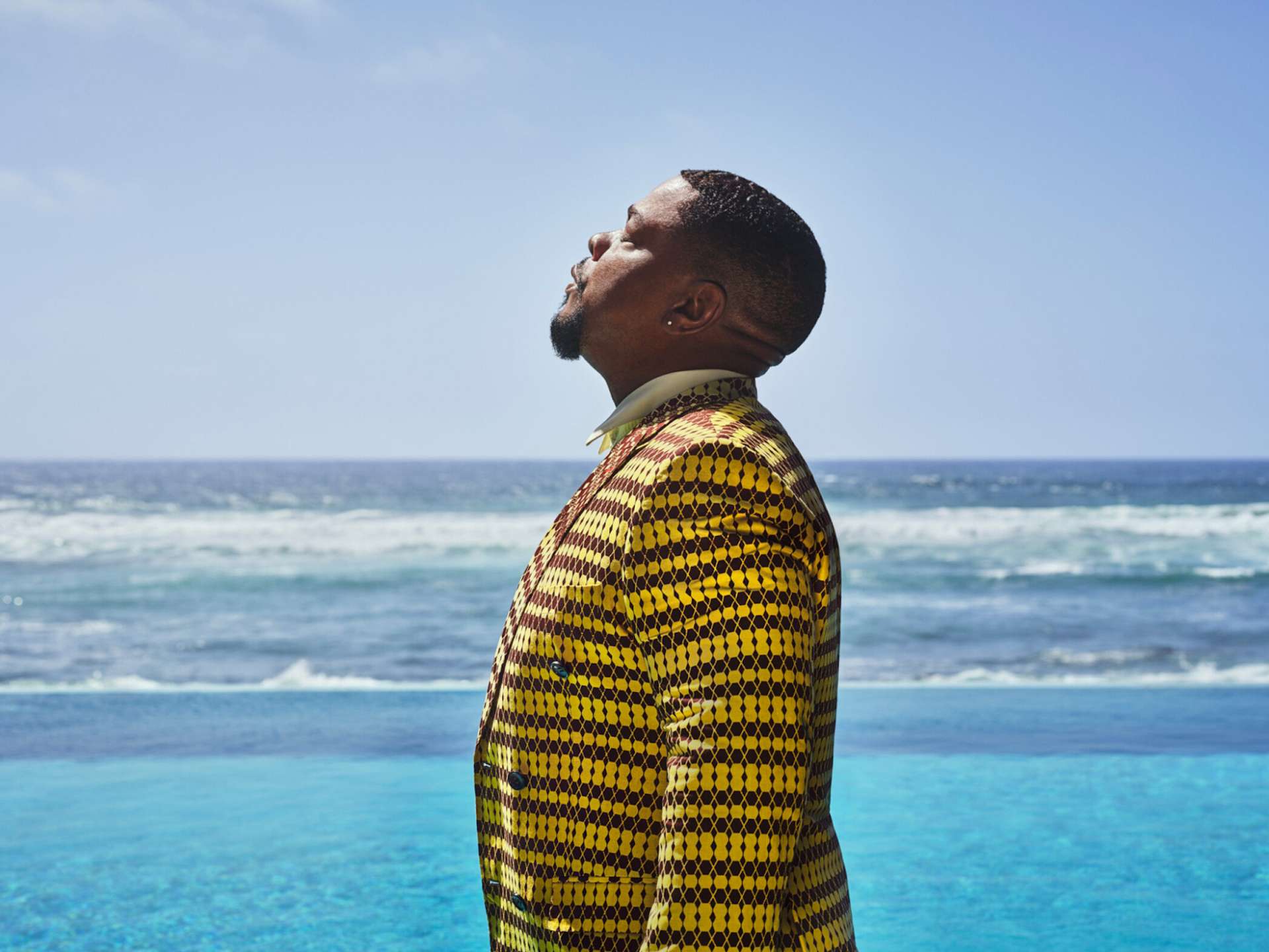Profile view of Kehinde Wiley at his artist residence in Dakar, Senegal. He wears a yellow patterned blazer and looks up to the sky with his eyes closed; behind him, an infinity pool merges into a panorama of the ocean.