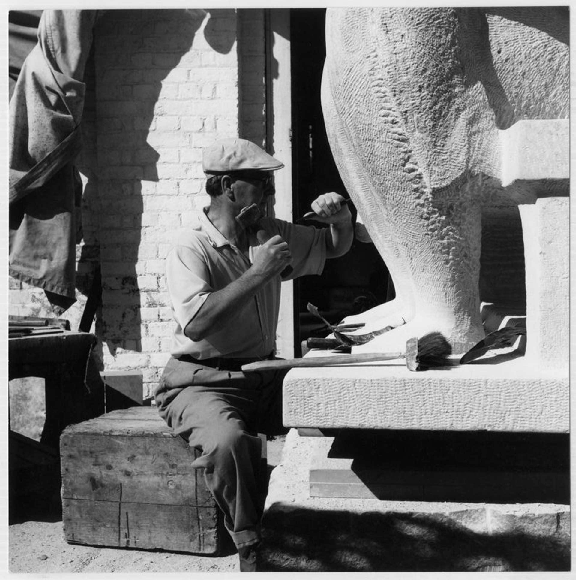 Black-and-white photograph of Henry Moore seated on a wooden plinth, wearing trousers, a white short-sleeve shirt, and a newsboy cap. Moore is seen carving at the foot of a large-scale sculpture, which extends beyond the frame of the photograph.