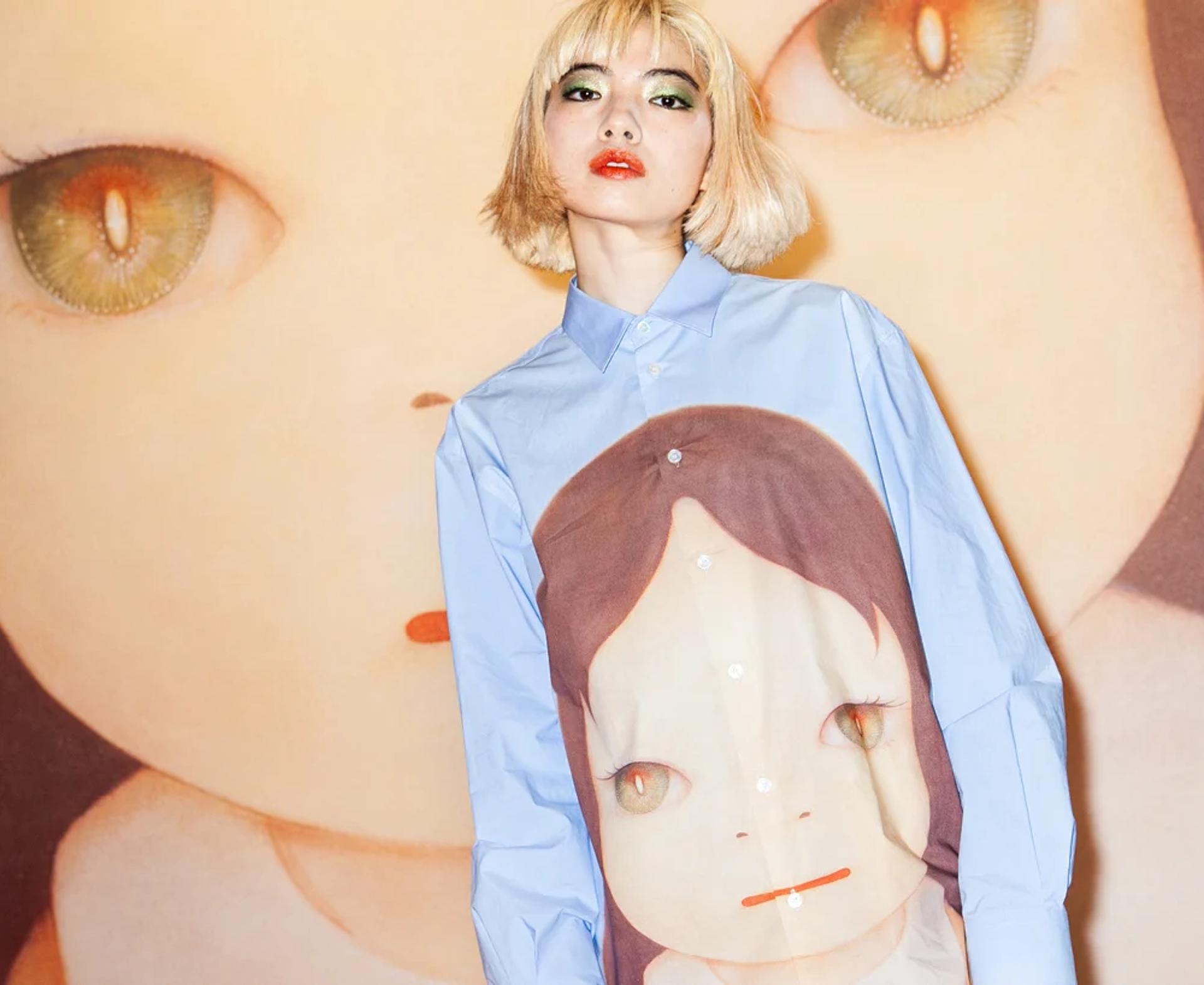 An image of a model wearing a shirt featuring artwork by the artist Yoshitomo Nara, standing against a background featuring that same artwork.
