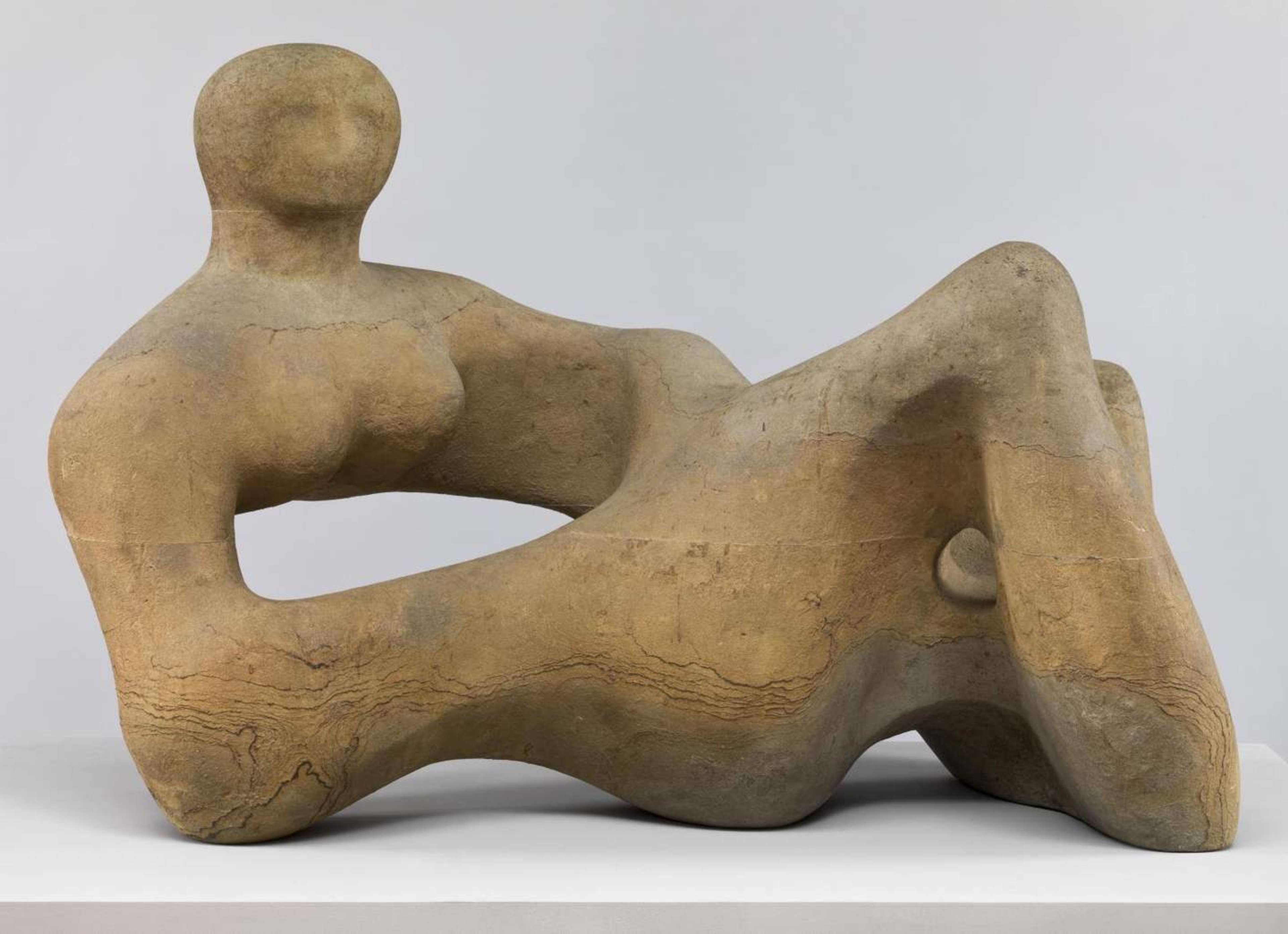 Abstract sculpture with exaggerated and distorted features. The reclining figure rests on its forearms, with a hollow hole in the centre of its chest.