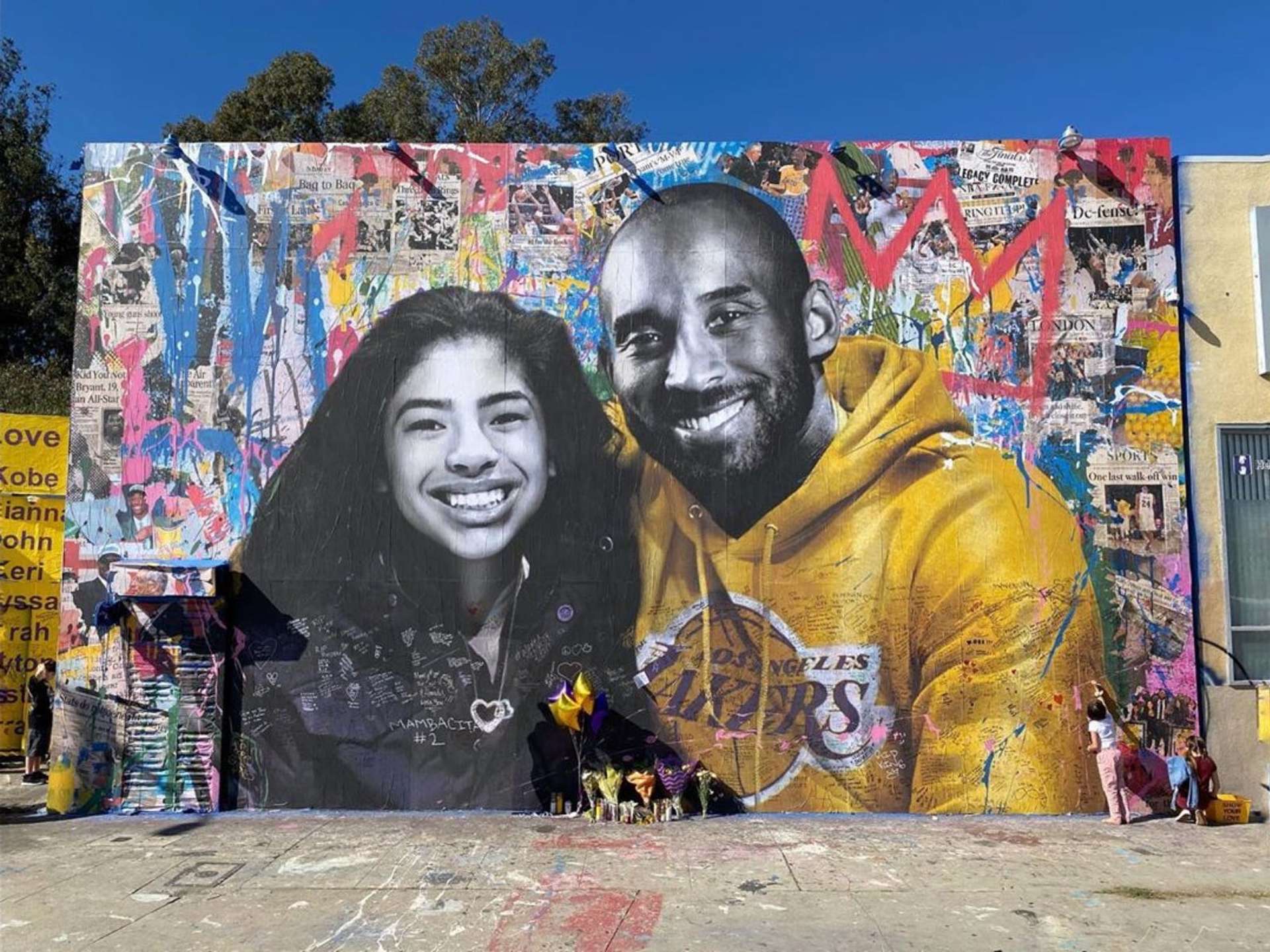 A photograph of the Kobe Bryant Mural by Mr. Brainwash, showing a monochrome image of athlete Kobe Bryant, wearing a bright yellow Los Angeles Lakers sweatshirt, and his daughter Gianna. They are posing against a background featuring a spray painted wall of several newspapers highlighting some of Kobe’s biggest achievements. In the corner behind Kobe is one of Basquiat’s crowns.