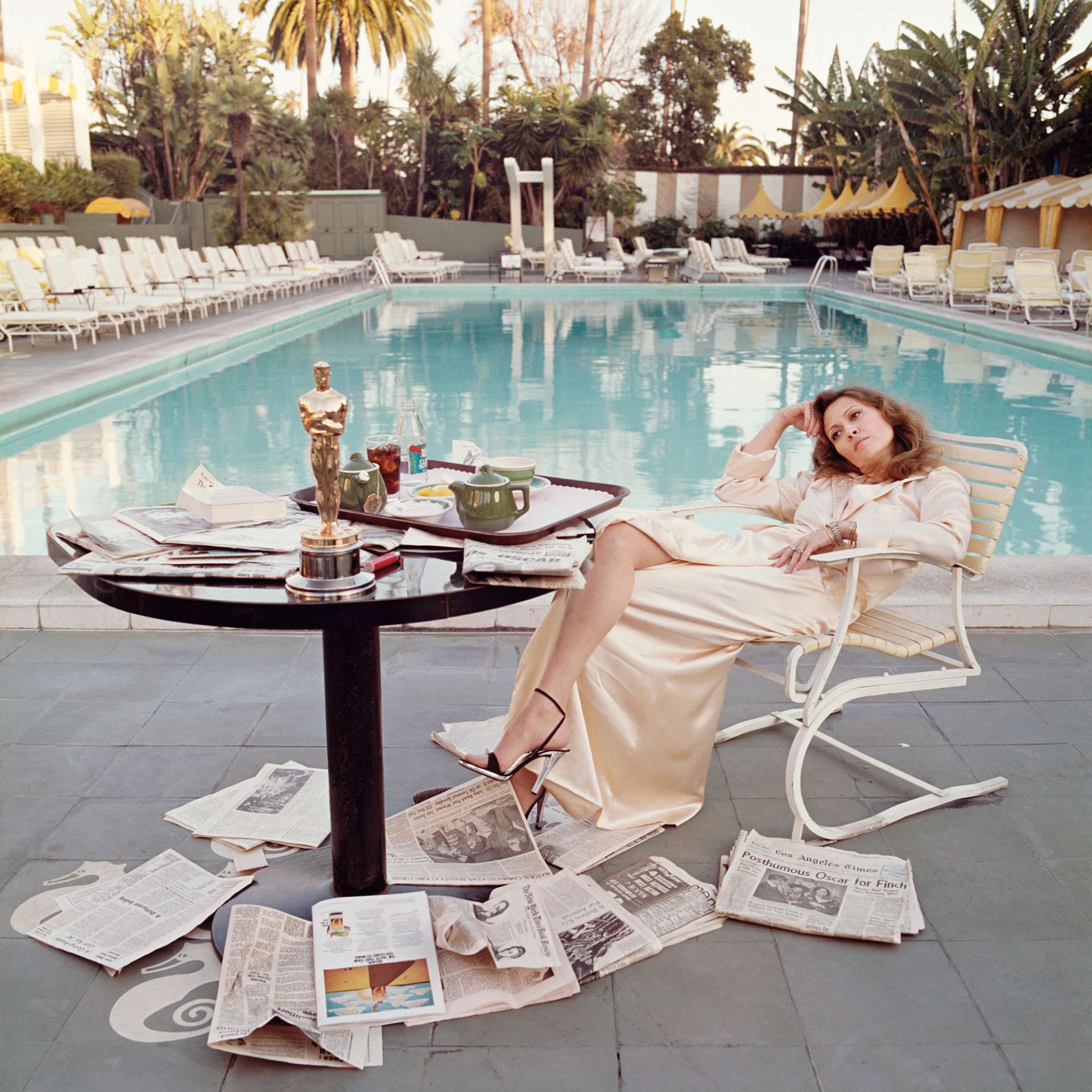 A colour photograph by Terry O'Neill depicting Faye Dunaway in a cream silk gown, lounging on a chair with a swimming pool in the background. On the table to the left of the frame is an Oscar award, a tray with a pot of tea, and newspapers which also litter the ground about her.