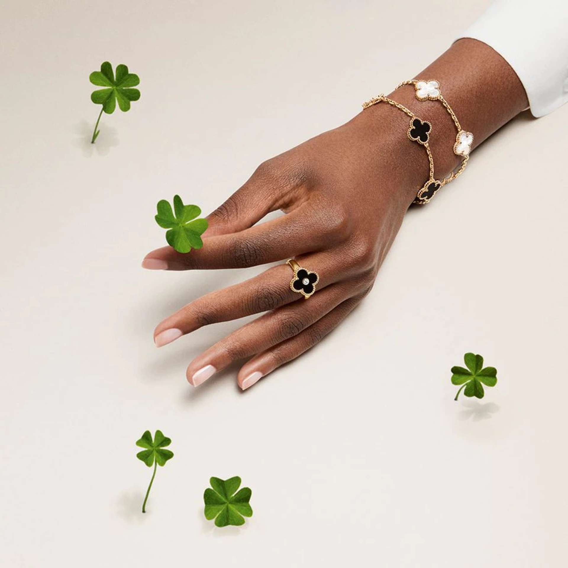 Image of a hand holding four leaf clover with 4 other clovers surrounding the hand, wearing a black clover ring and two bracelets - one gold and black, and the other pearl and gold