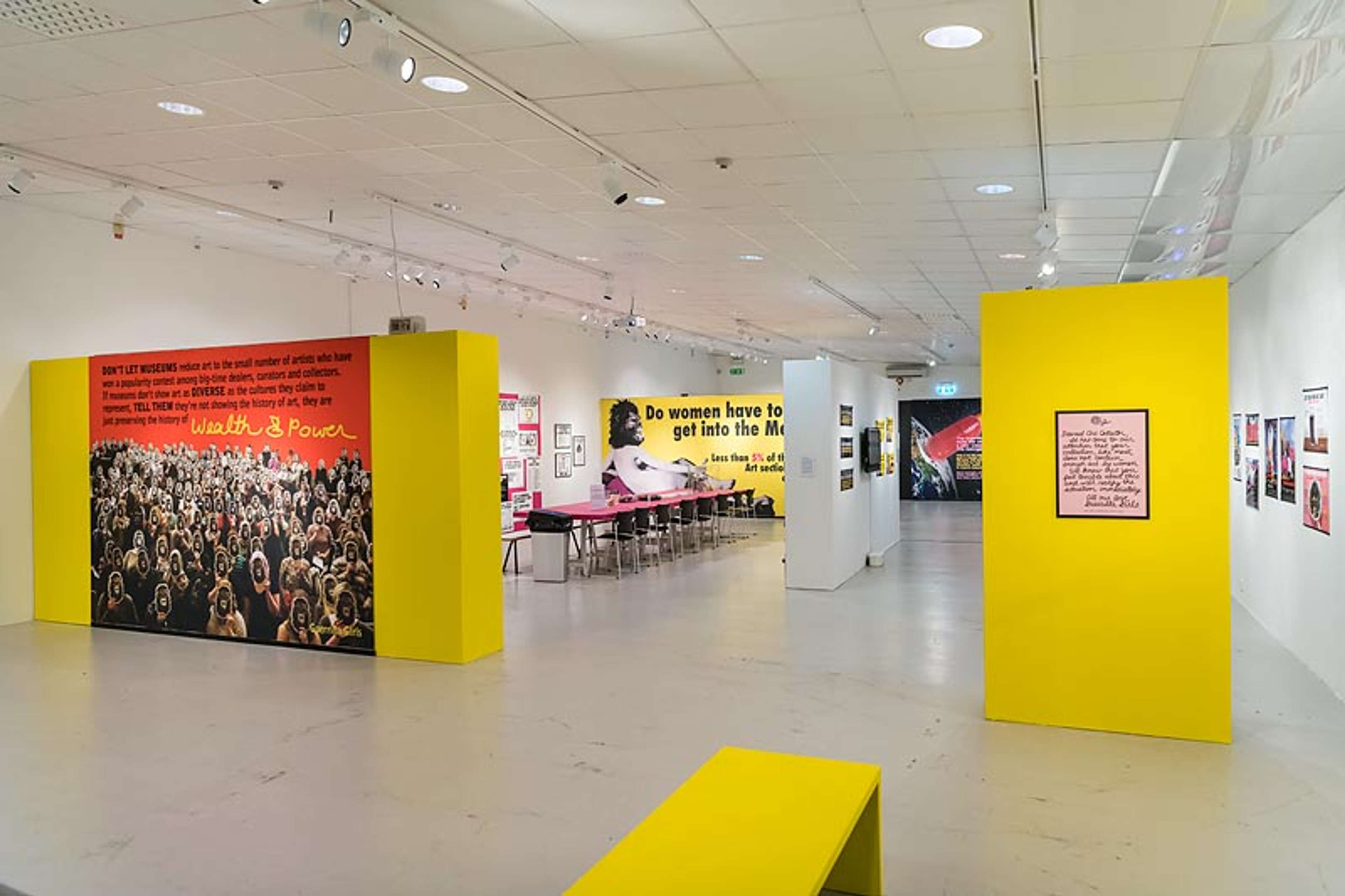 A contemporary art exhibition featuring Guerrilla Girls' activist artwork. The space includes bold yellow display panels, feminist protest posters, and a large banner reading, "Do women have to be naked to get into the Met Museum?" The exhibit critiques gender inequality in the art world through visually striking and provocative messages.