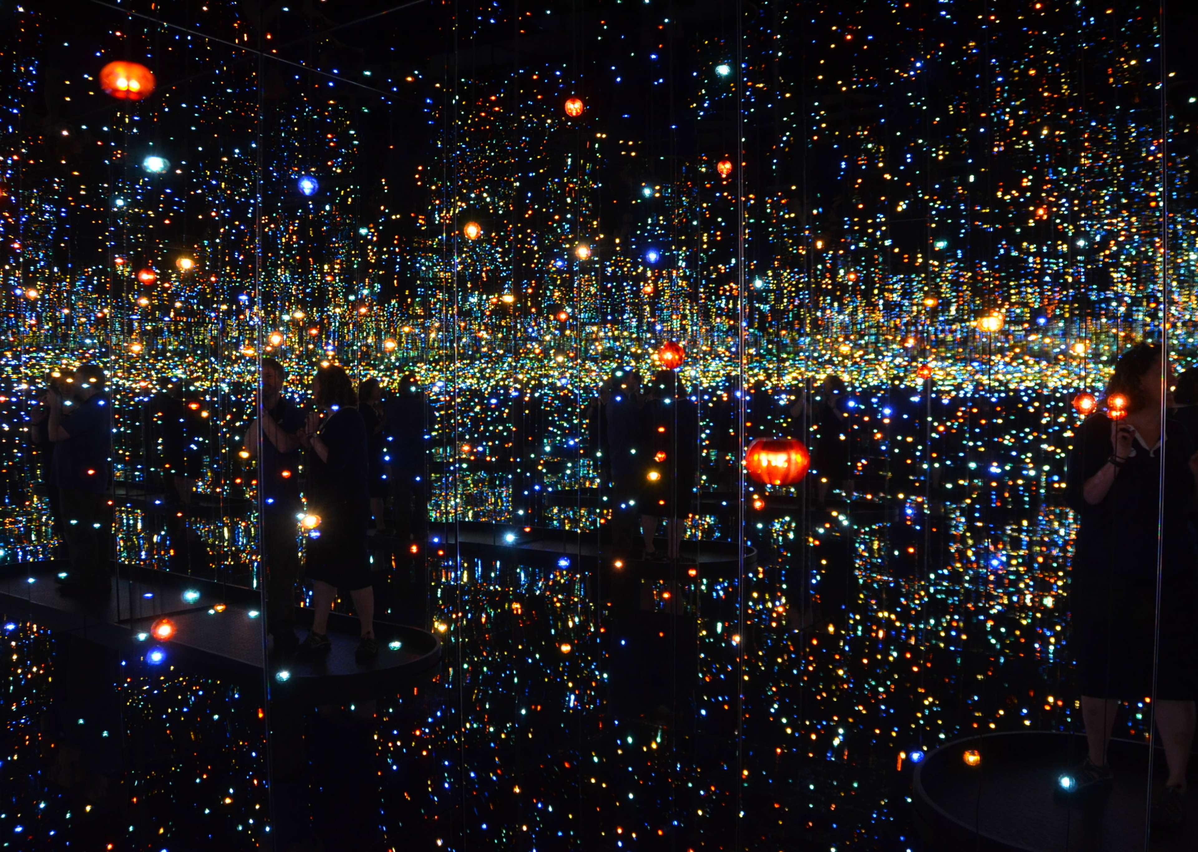 A photograph depicting a dark room lined with mirrors and filled with countless lights and lanterns in many colours. Dark figures of people walking around the room are reflected in the mirrors also and can be seen looking at the many lights.