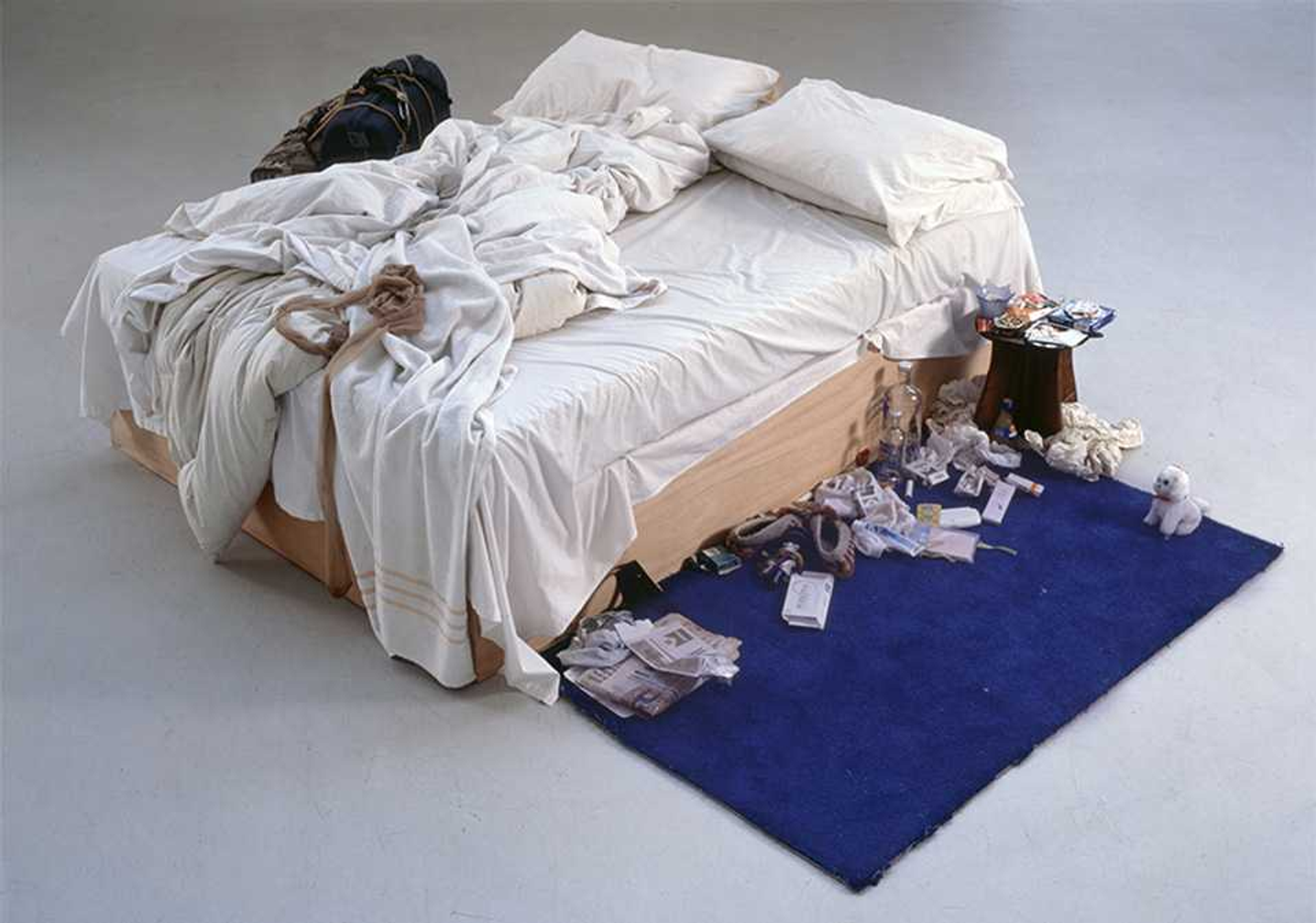 Photograph of Tracey Emin’s My Bed. An unmade bed with bunched up bedding, next to a blue rug covered with junk and personal items.