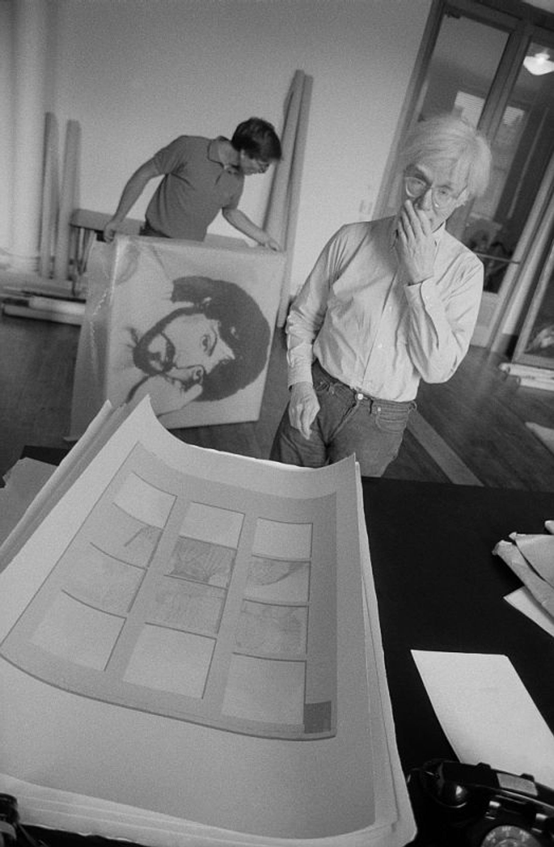 A black and white image of Andy Warhol looking at a silkscreen of banana split by Thomas Dellert.