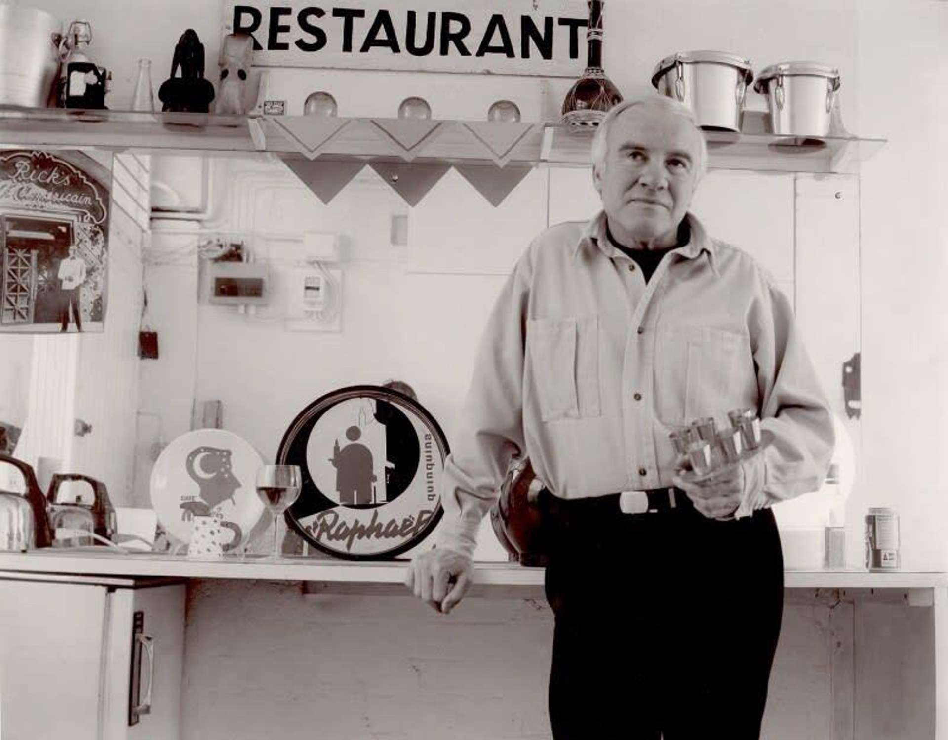 A black and white image of the artist Patrick Caulfield, standing in front of a kitchen.