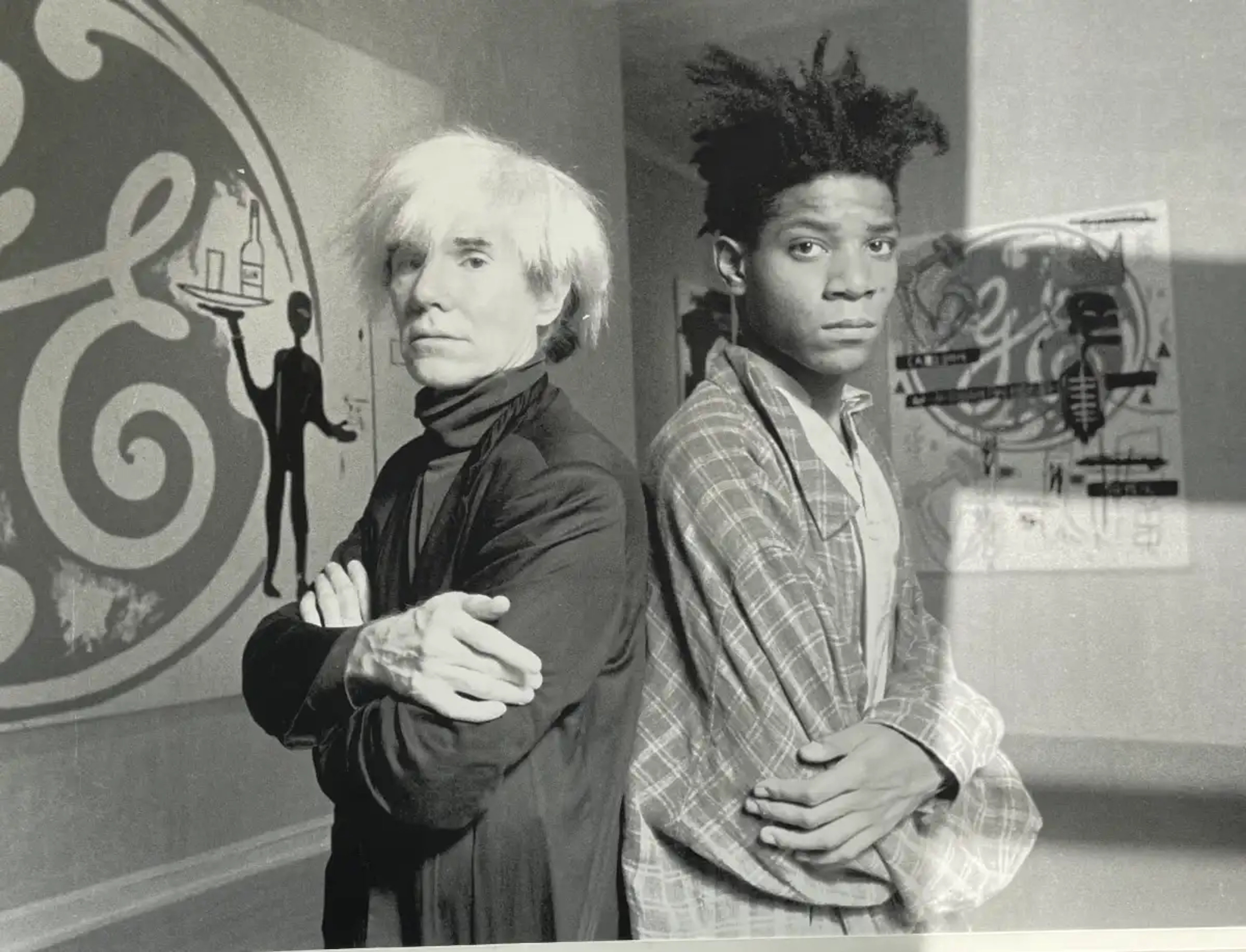 A monochrome photograph of Andy Warhol and Jean-Michel Basquiat, with their backs turned to one another, as they stand in front of several of the paintings they co-created.