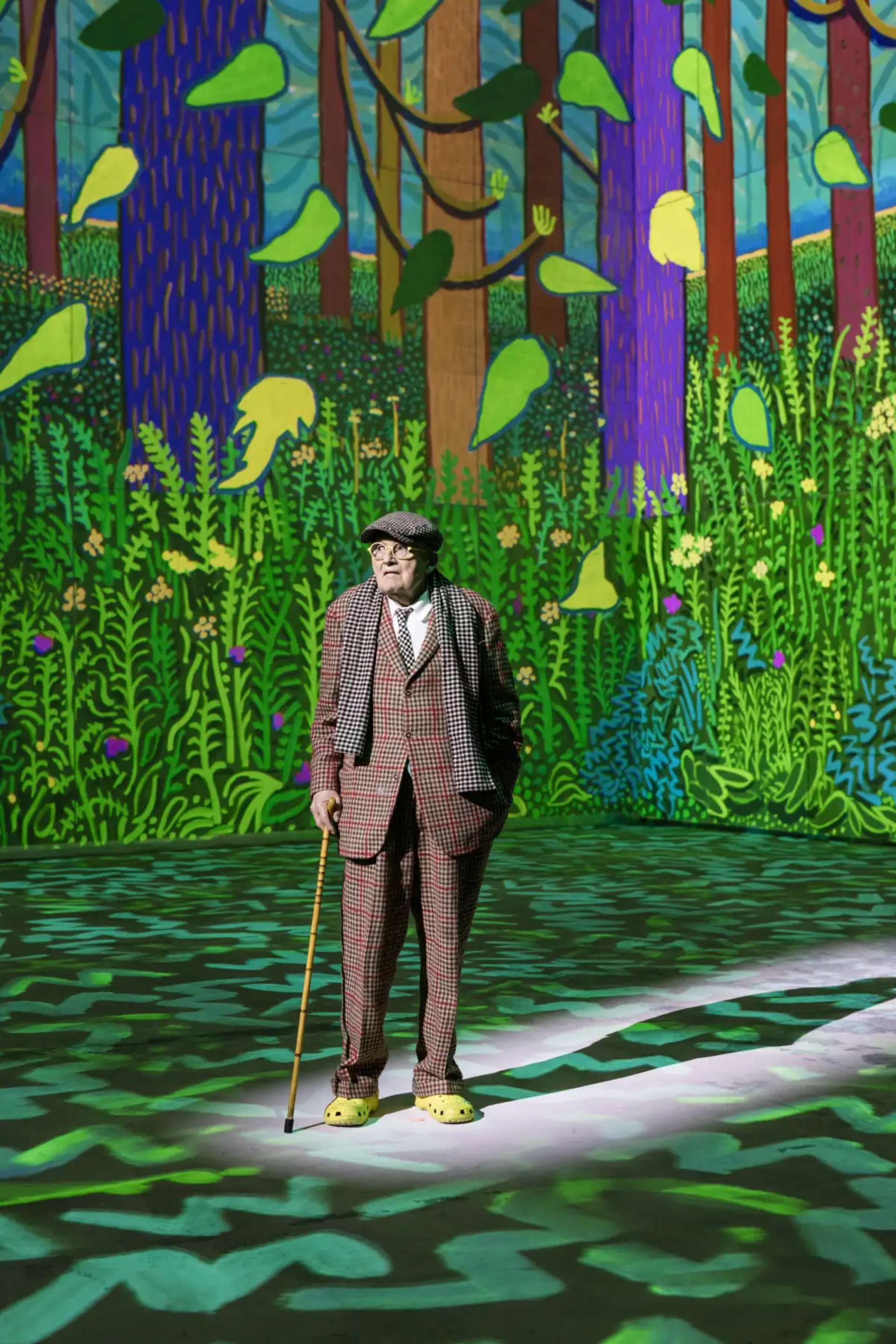 A photograph of artist David Hockney wearing a plaid suit and yellow Crocs in his Lightroom exhibition, with his digital drawing of a green forest projected on the walls and floor about him.